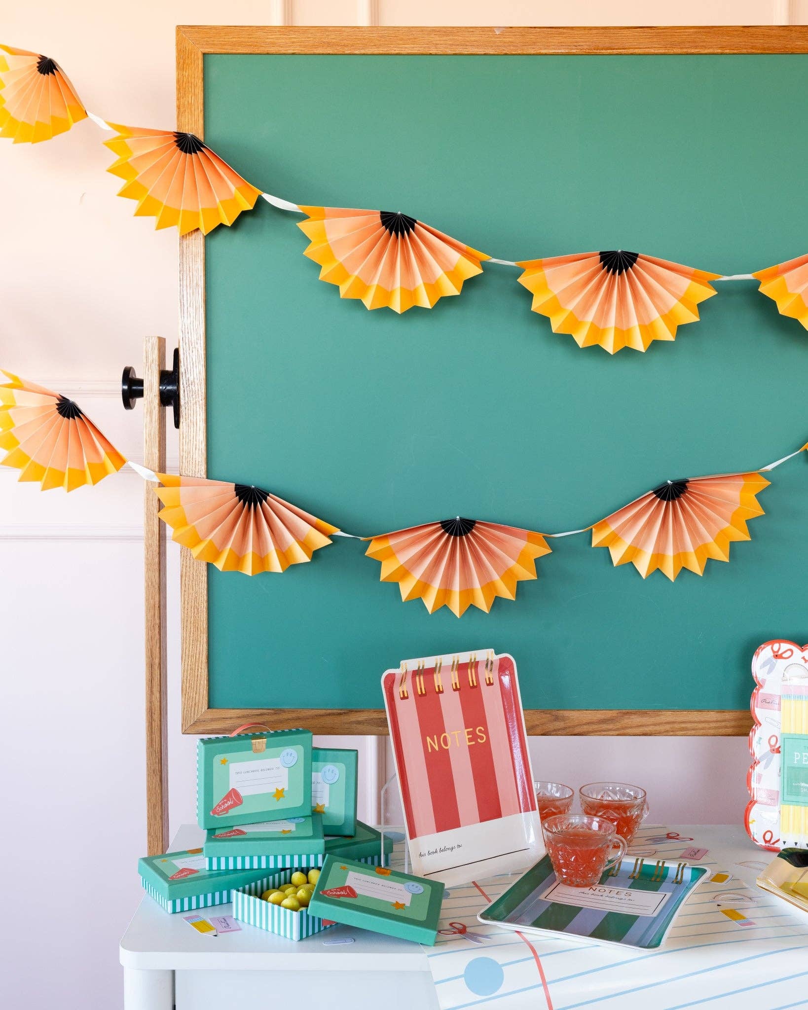 Close-up of two pencil shaving accordion banners hanging in tiers against green chalkboard with notebook-style table and striped notebook decor
