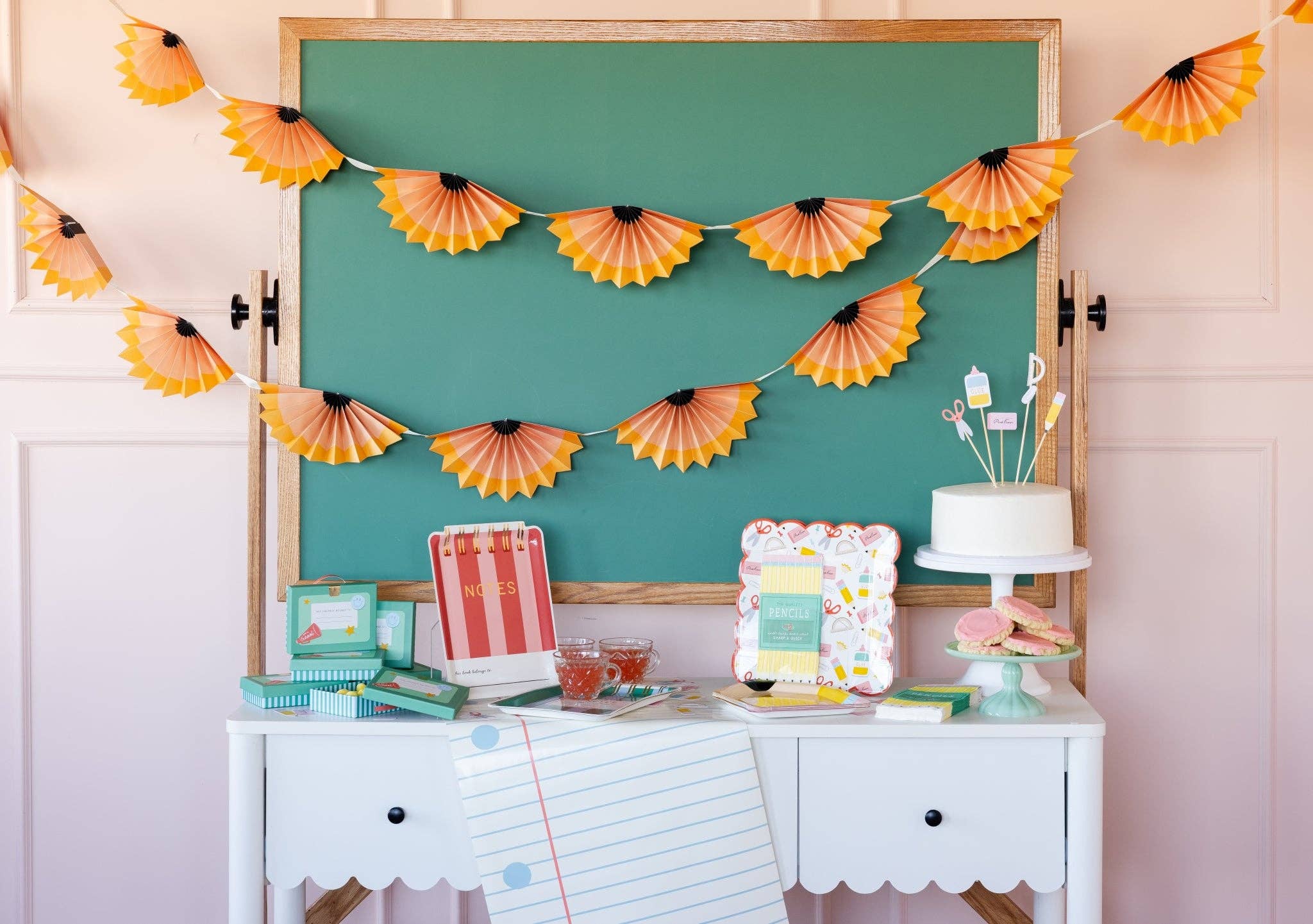 Styled classroom party with chalkboard backdrop, two strands of orange pencil shaving banner, notebook table runner, and striped "Notes" notebook on white desk