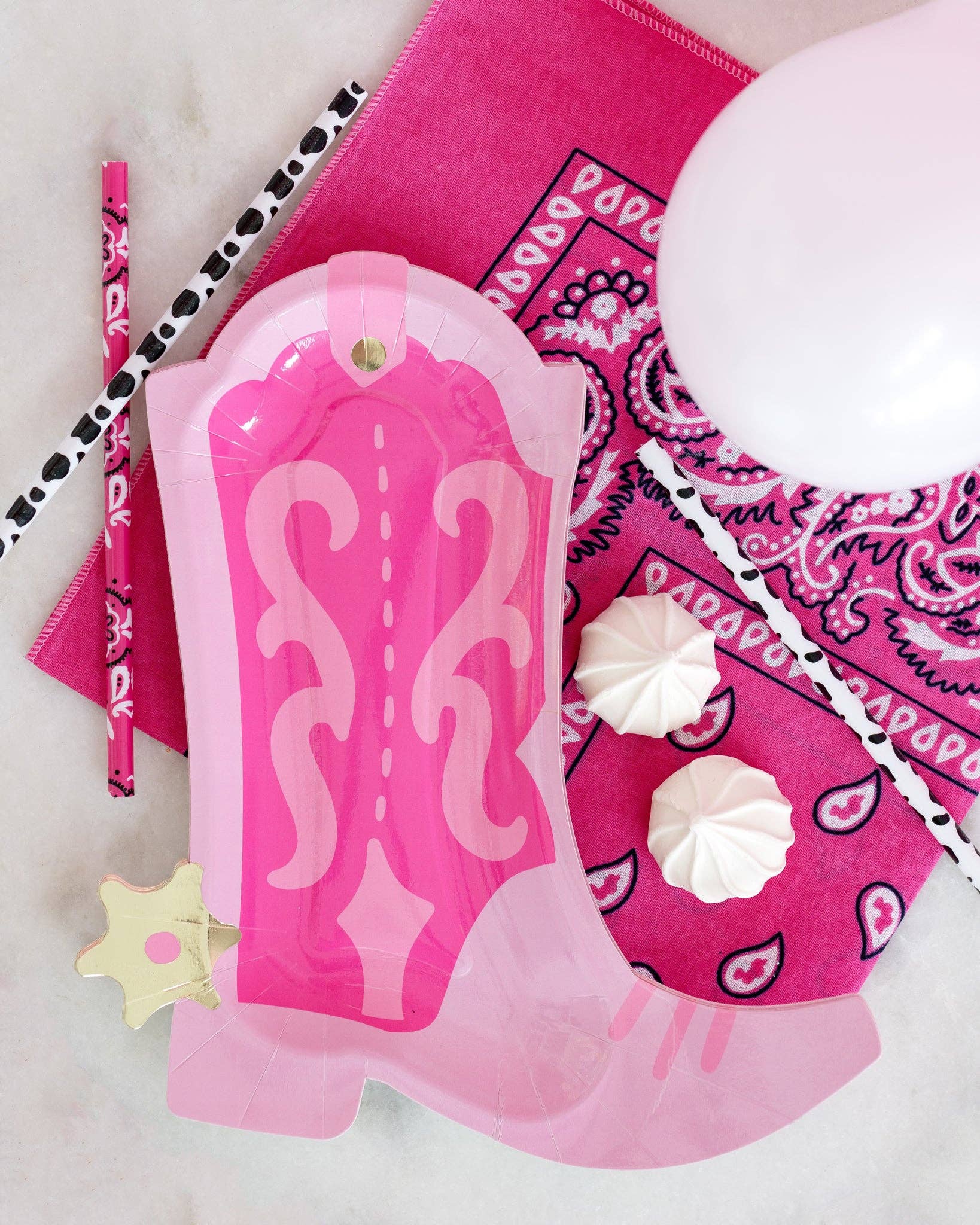 Pink cowgirl boot paper plate styled flat lay on hot pink bandana with cow print straws, pink paisley straws, pink balloon, and meringue cookies for a Nashville bachelorette tablescape