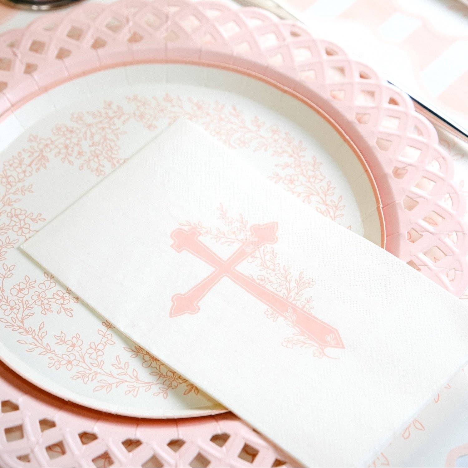 A close up of a soft pink lattice-edge paper plate with a storybook religious pink cross napkin for a baby dedication.