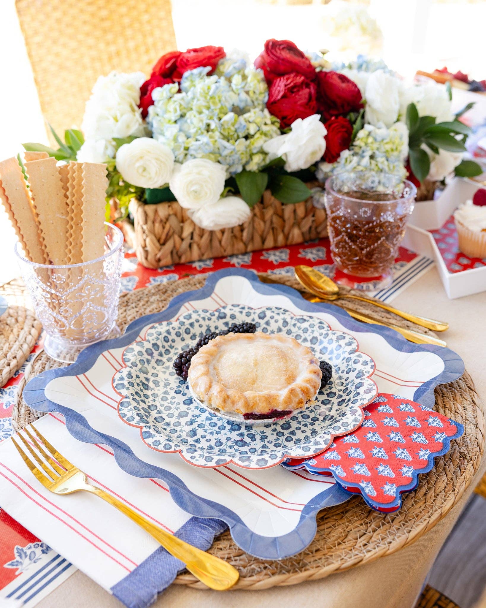 Plaid stars scalloped paper plate as charger under blue ditsy floral plate with mini pie, red block-print napkin, wicker placemat, and gold cutlery in Hamptons Americana setting