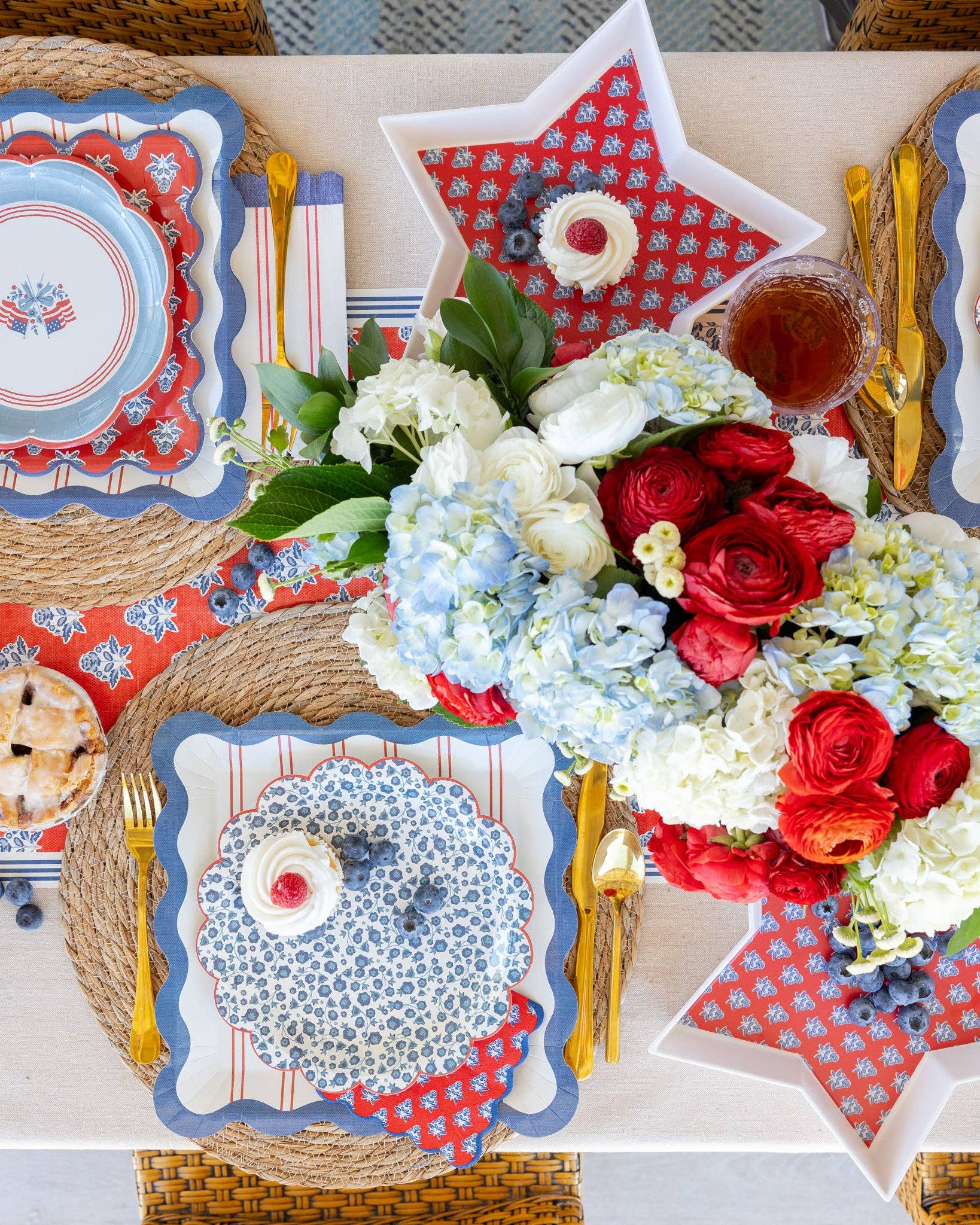 Hamptons Americana full tablescape overhead with plaid star plates, blue ditsy floral plates, American flag plates, red block-print napkins, star trays, hydrangeas, and gold cutlery