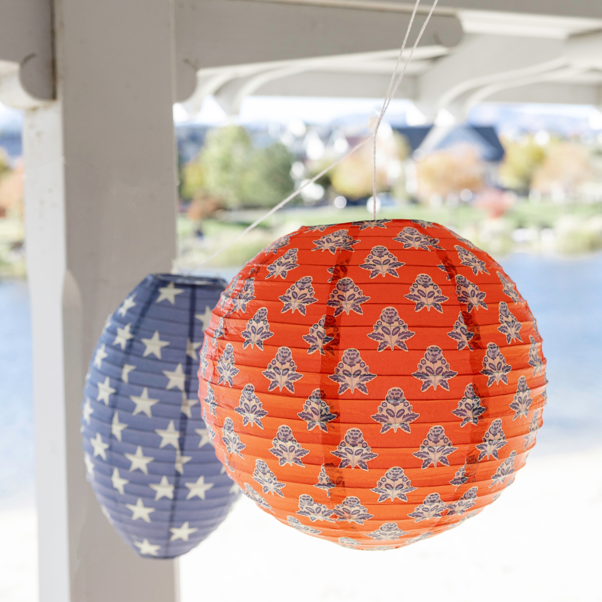 Red block print floral paper lantern and navy star paper lantern hanging from white porch beam with lake view behind