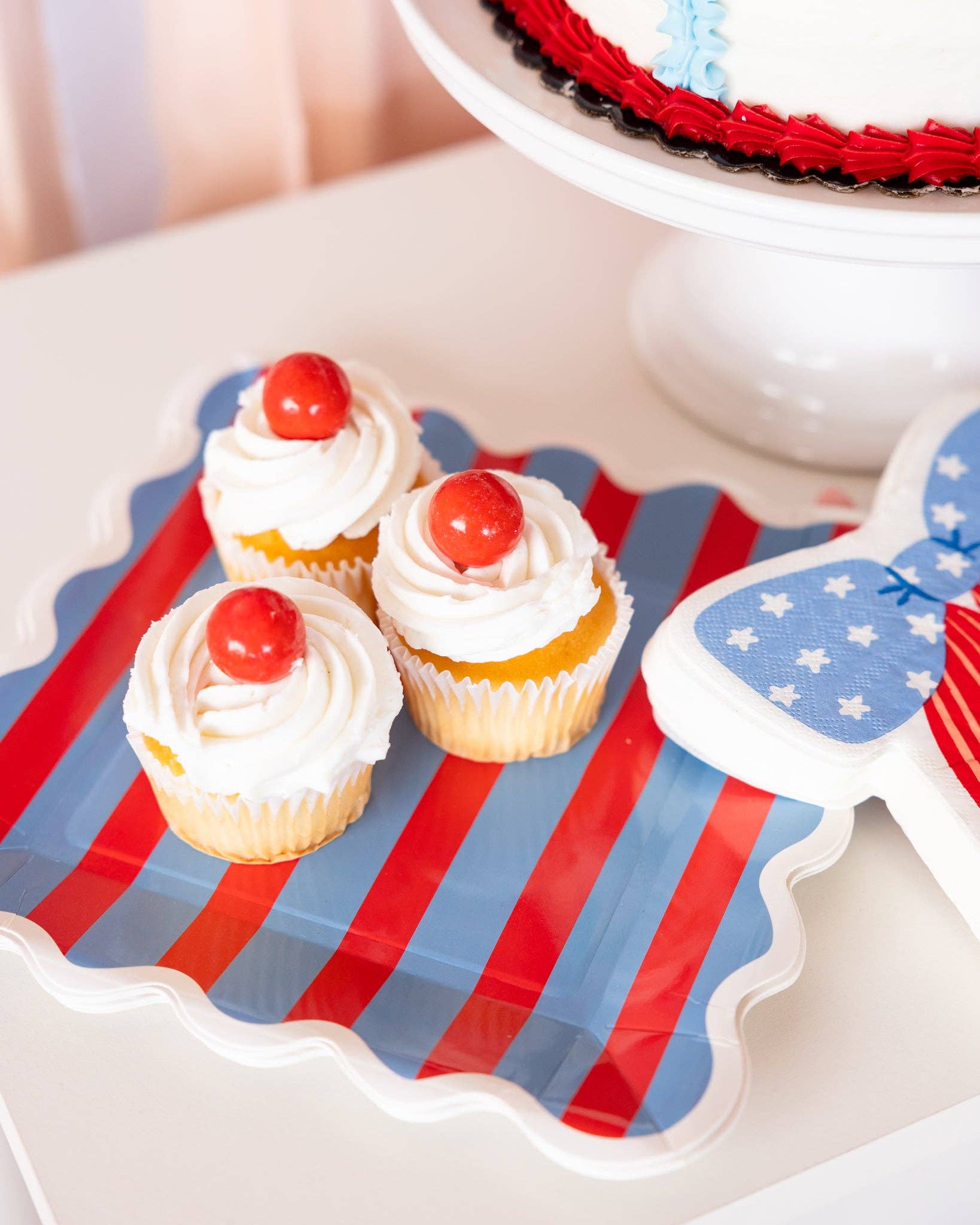 Overhead close-up of cherry-topped cupcakes on a red and blue striped scalloped paper plate next to a bow shaped napkin and cake stand