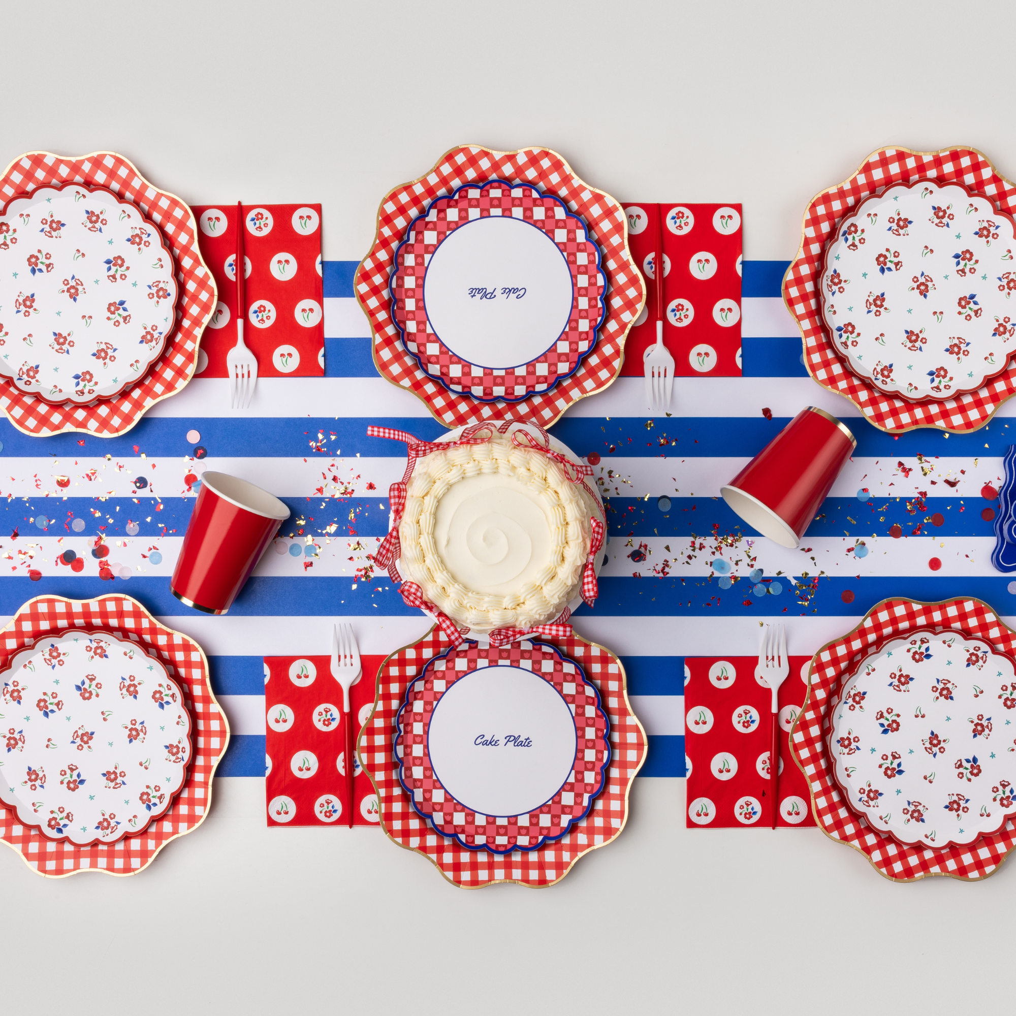 Overhead styled table with red gingham cake plates, floral plates, cherry print napkins, red polka dot celebrate napkins, red cups, confetti, and cake on blue striped table runner