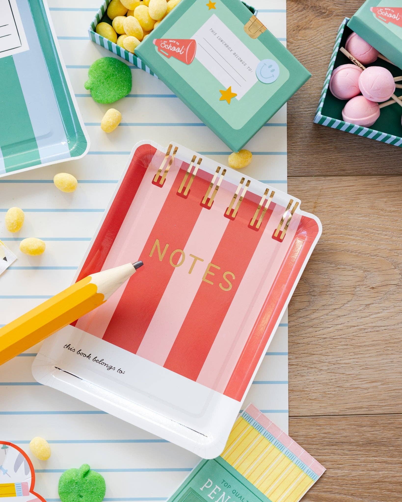 Flat lay of red and pink striped notebook shaped paper plate with oversized paper pencil prop, green Back to School lunchbox treat boxes, and pencil napkins