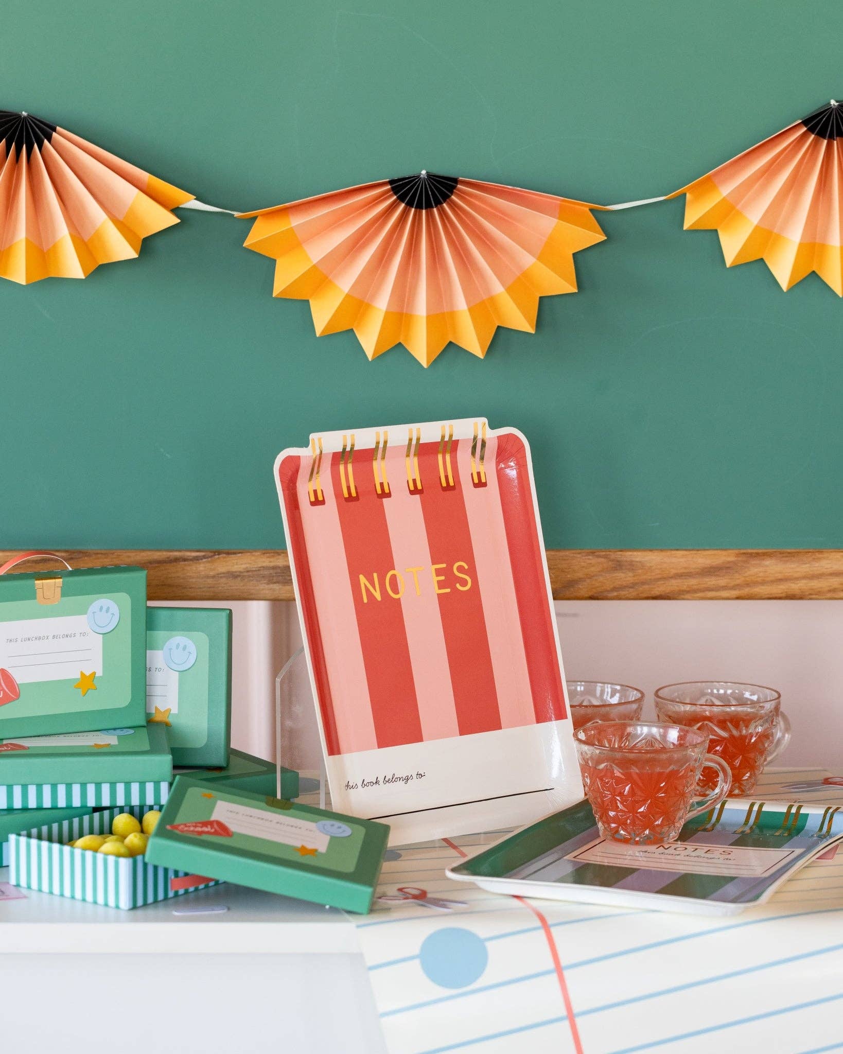 Red and pink striped notebook shaped paper plate displayed upright on an acrylic stand against a green chalkboard wall with pencil shaving accordion banner and school-themed treat boxes