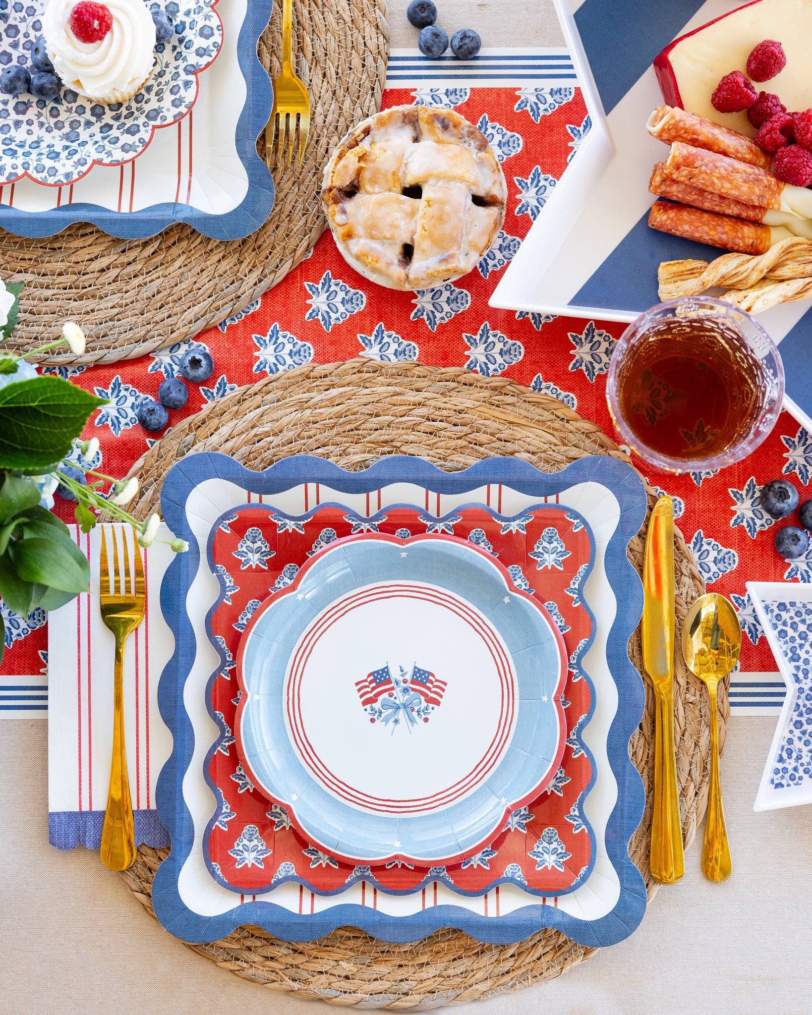 Overhead Americana place setting with layered scalloped plates, red striped dinner napkin, and gold flatware on woven charger