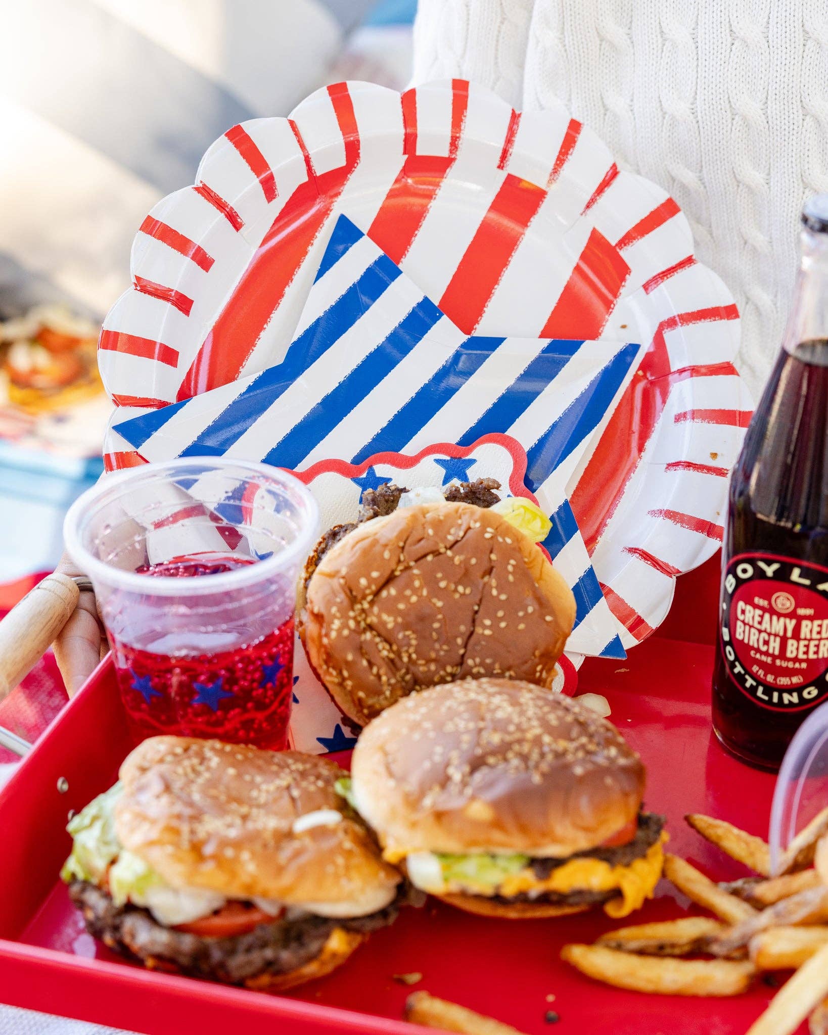 Red striped scalloped paper dinner plate with blue striped napkin on a red serving tray with burgers, fries, and a patriotic cup