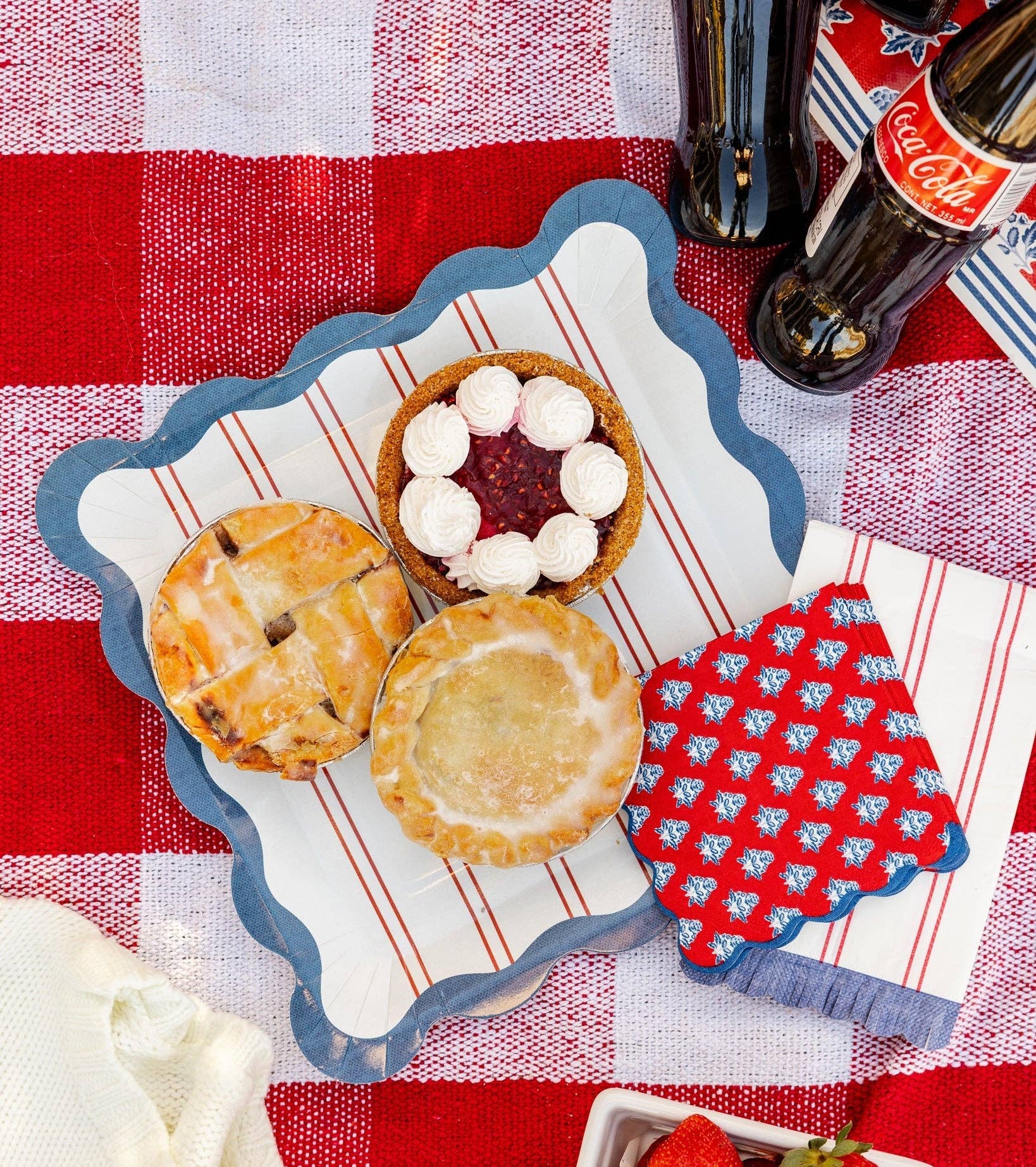 Red striped scalloped paper plate with mini pies and red block-print floral napkin on red and white gingham picnic blanket — summer Americana styling