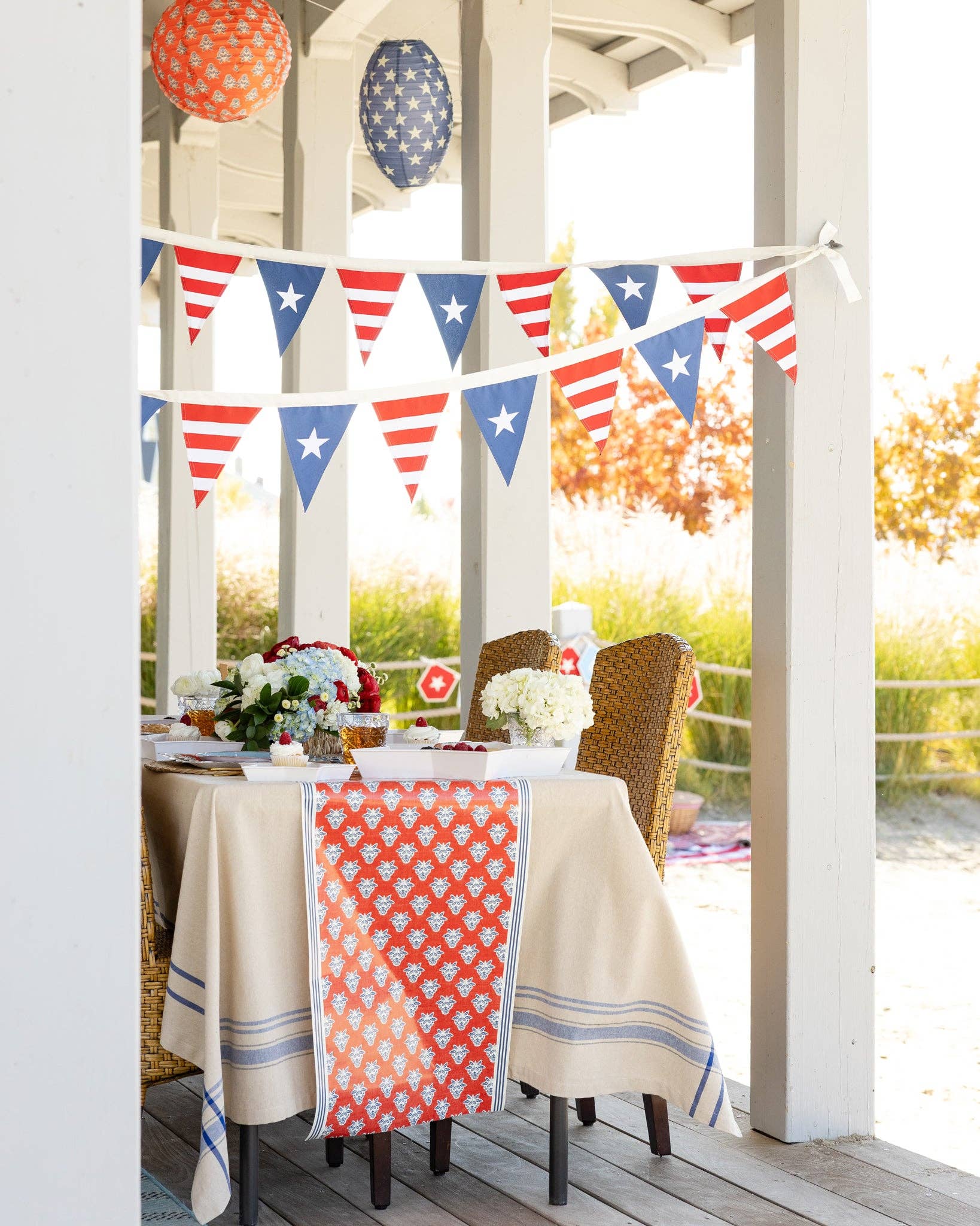Porch dinner table with red block print table runner draped over linen tablecloth with patriotic pennant bunting and paper lanterns
