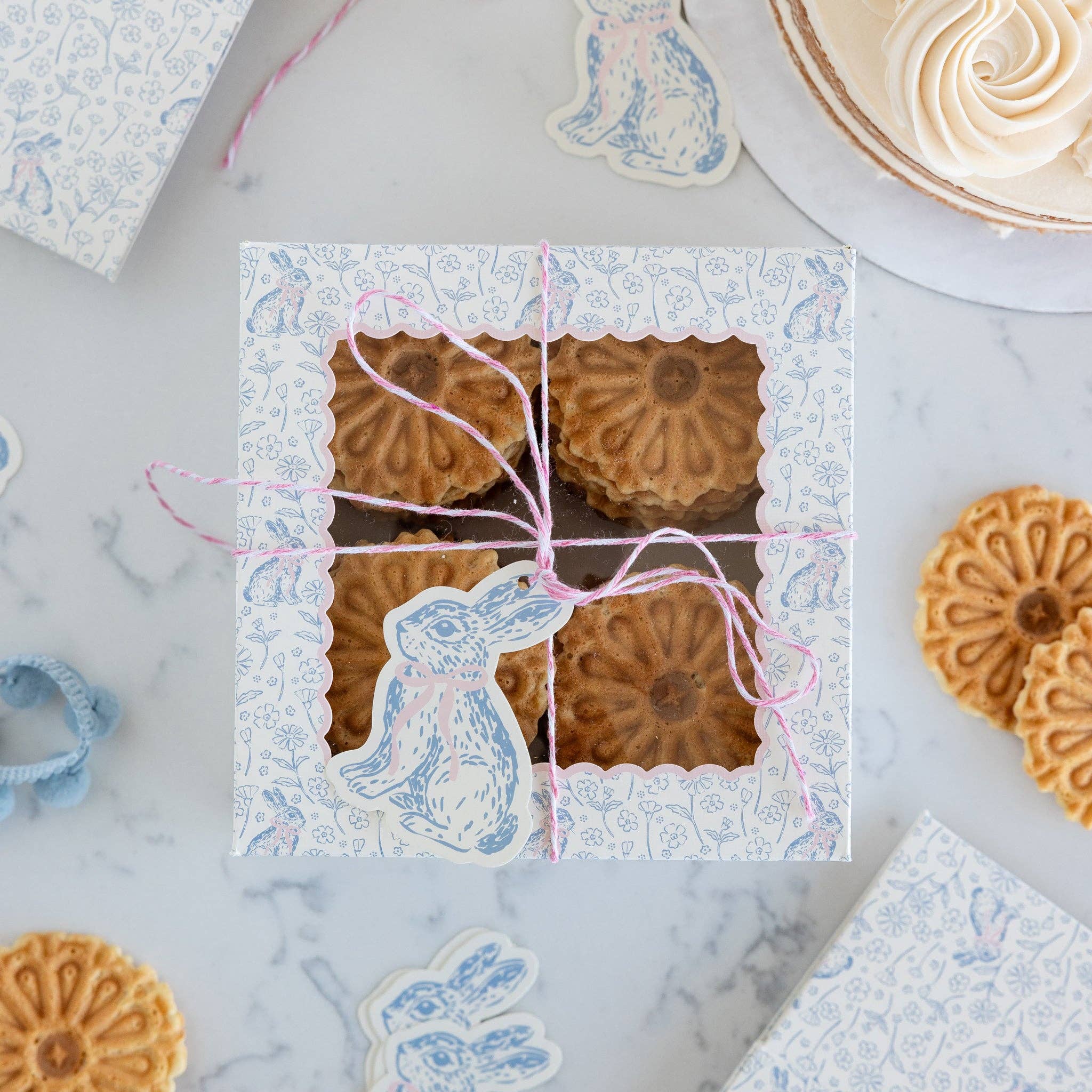 An overhead view of a blue-and-white floral treat box filled with flower-shaped cookies, tied with pink twine and a bunny tag for an Easter garden party.