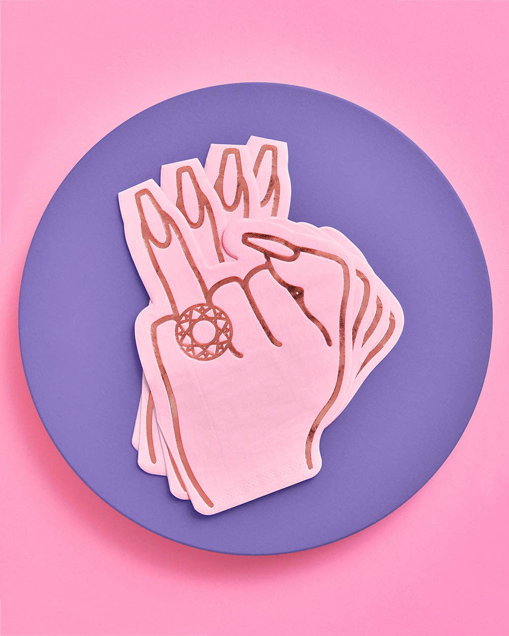 Stack of pink ring finger die-cut napkins on purple plate against pink background showing hand with diamond ring in hot pink foil