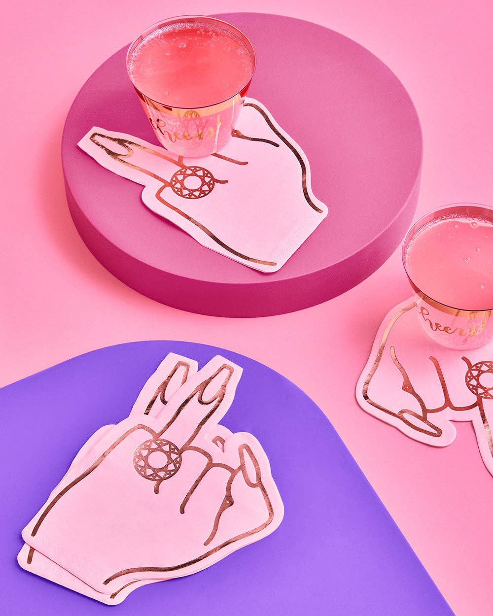 Pink ring finger napkins in two hand poses tucked under cocktail glasses on pink and purple surfaces at a bachelorette party setting