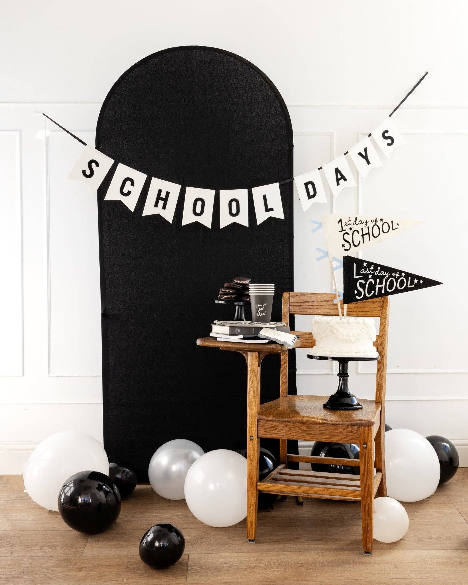School Days felt banner hung across a black arch backdrop with black and white balloons, wooden school chair, white layer cake, and 1st Day of School and Last Day of School pennants