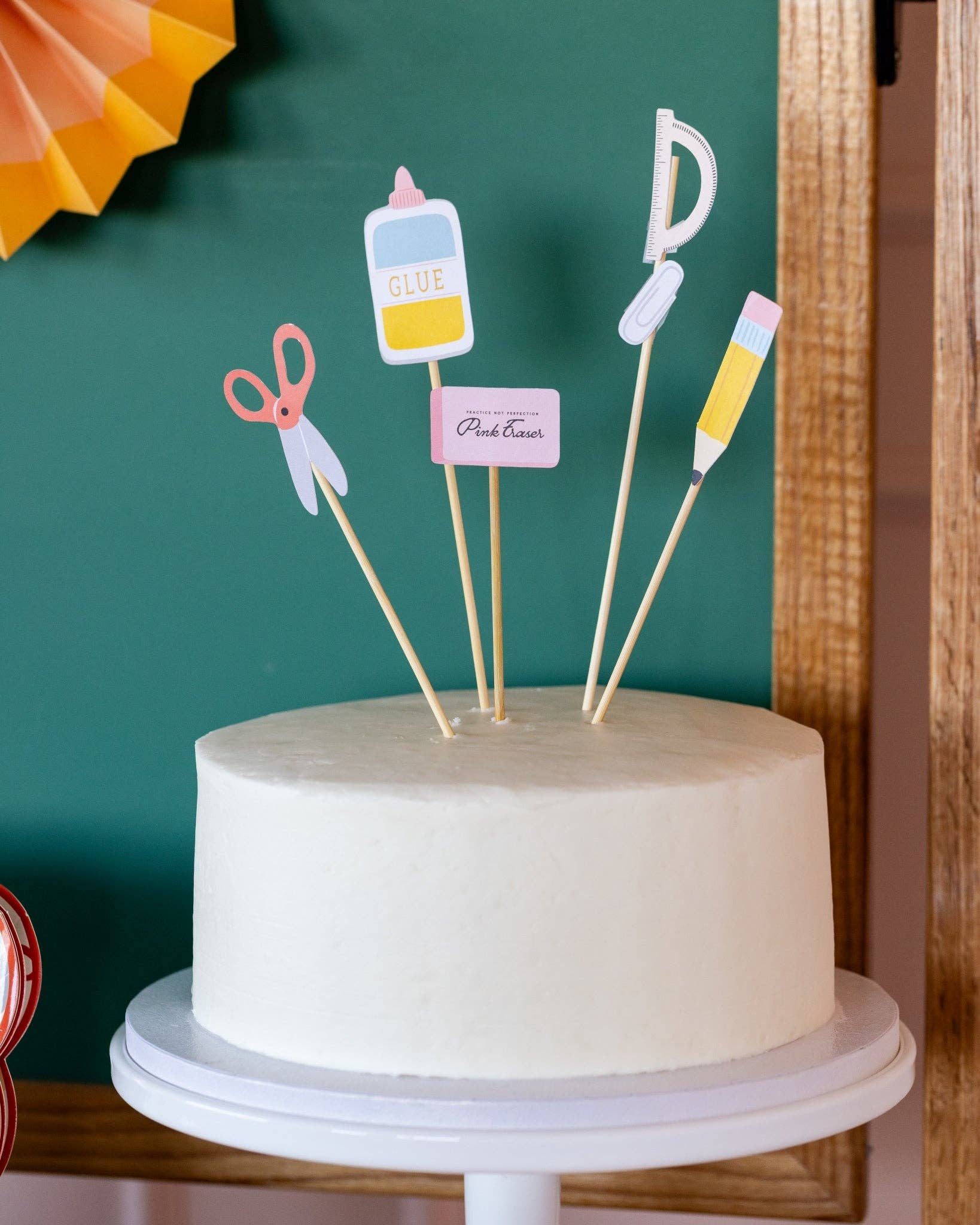 White cake styled with school supply paper picks including scissors, glue, pink eraser, protractor, paper clip, and pencil, against chalkboard backdrop with pencil shaving banner