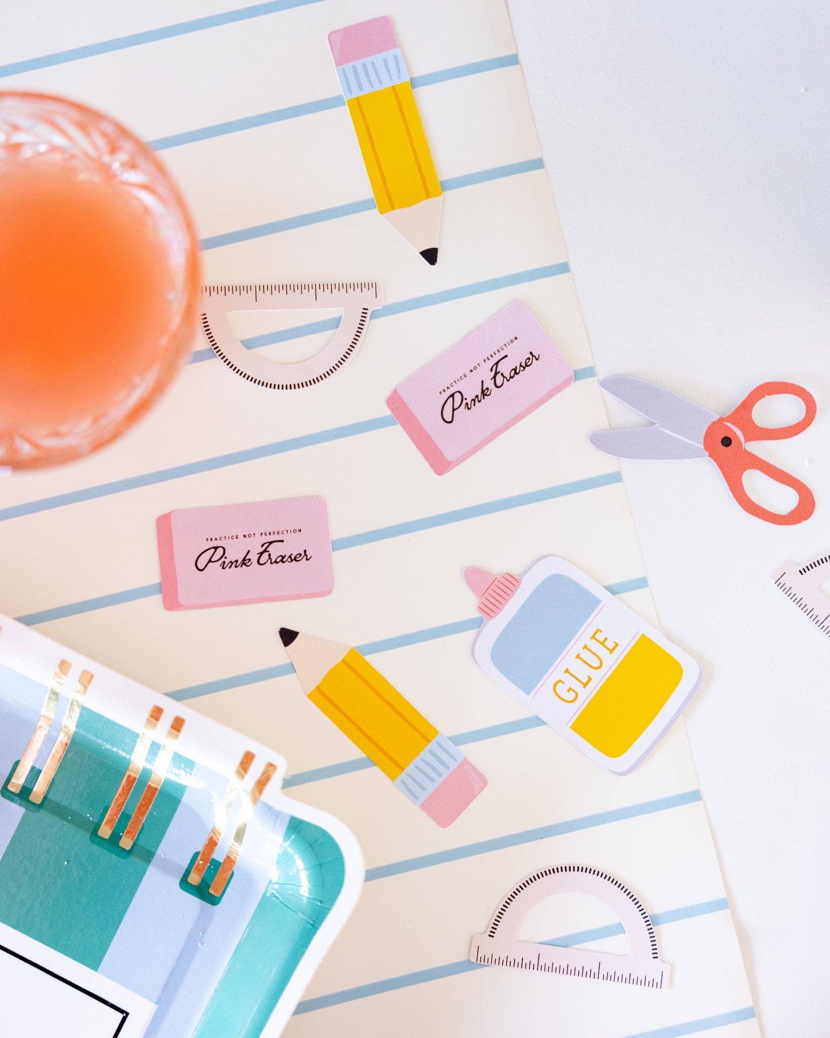 School supplies paper confetti pieces scattered on notebook paper background including pencils, pink erasers, protractors, scissors, and glue bottle, styled with orange juice and blue party plate