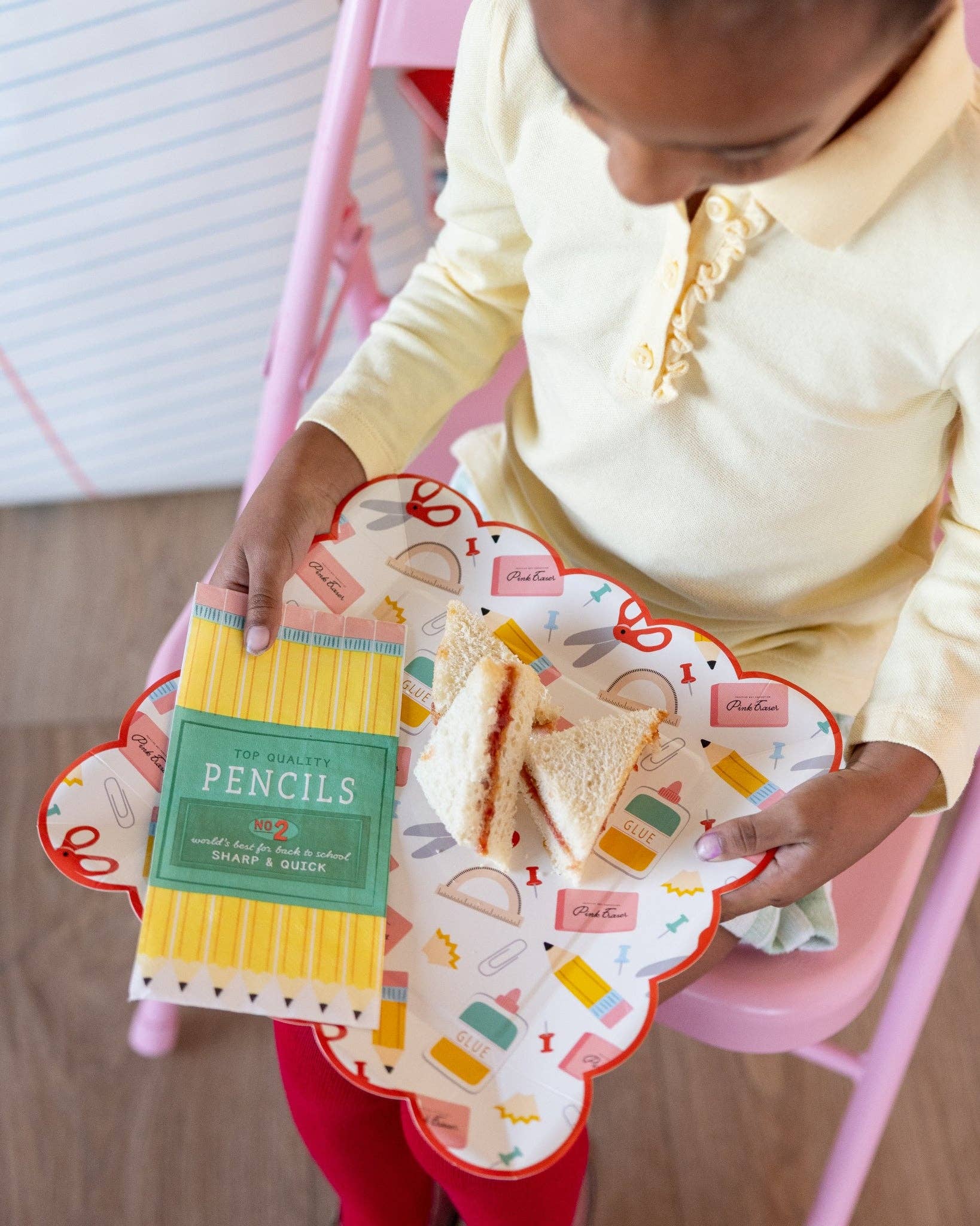 Young girl in yellow polo holding school supplies scalloped paper plate with PB&J sandwich and pencil box paper napkin