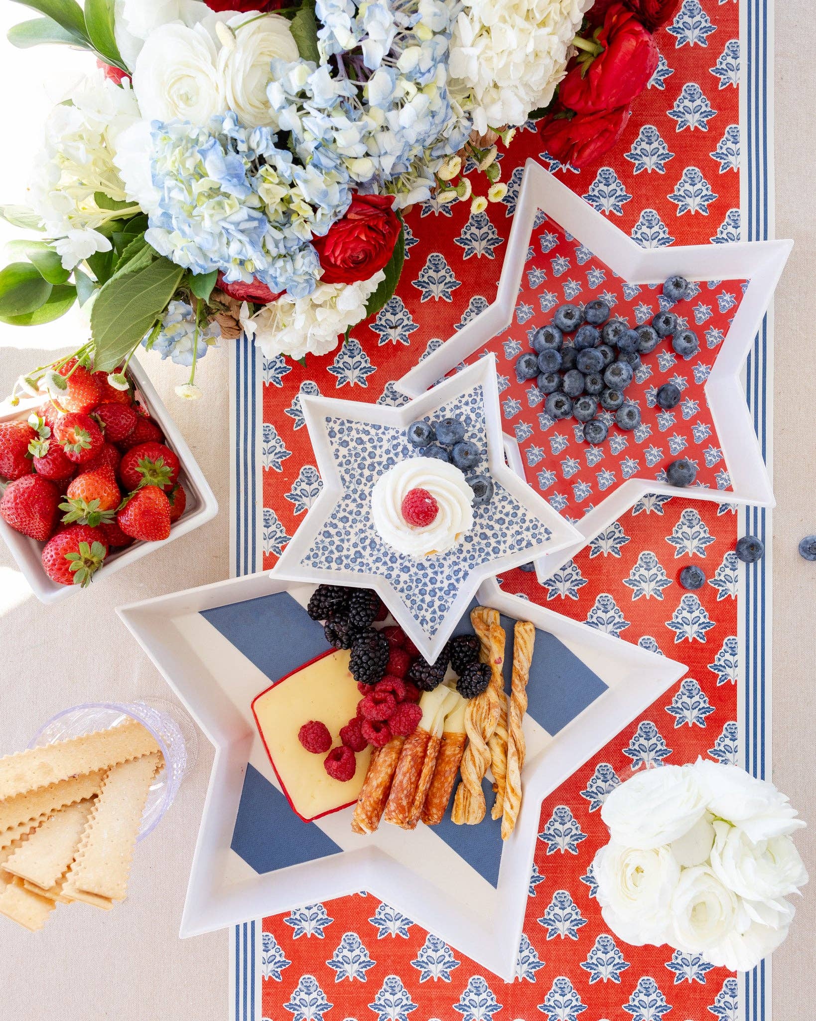 Three nesting star bamboo trays stacked with blueberries, cheese, breadsticks, and cupcake on red block print table runner