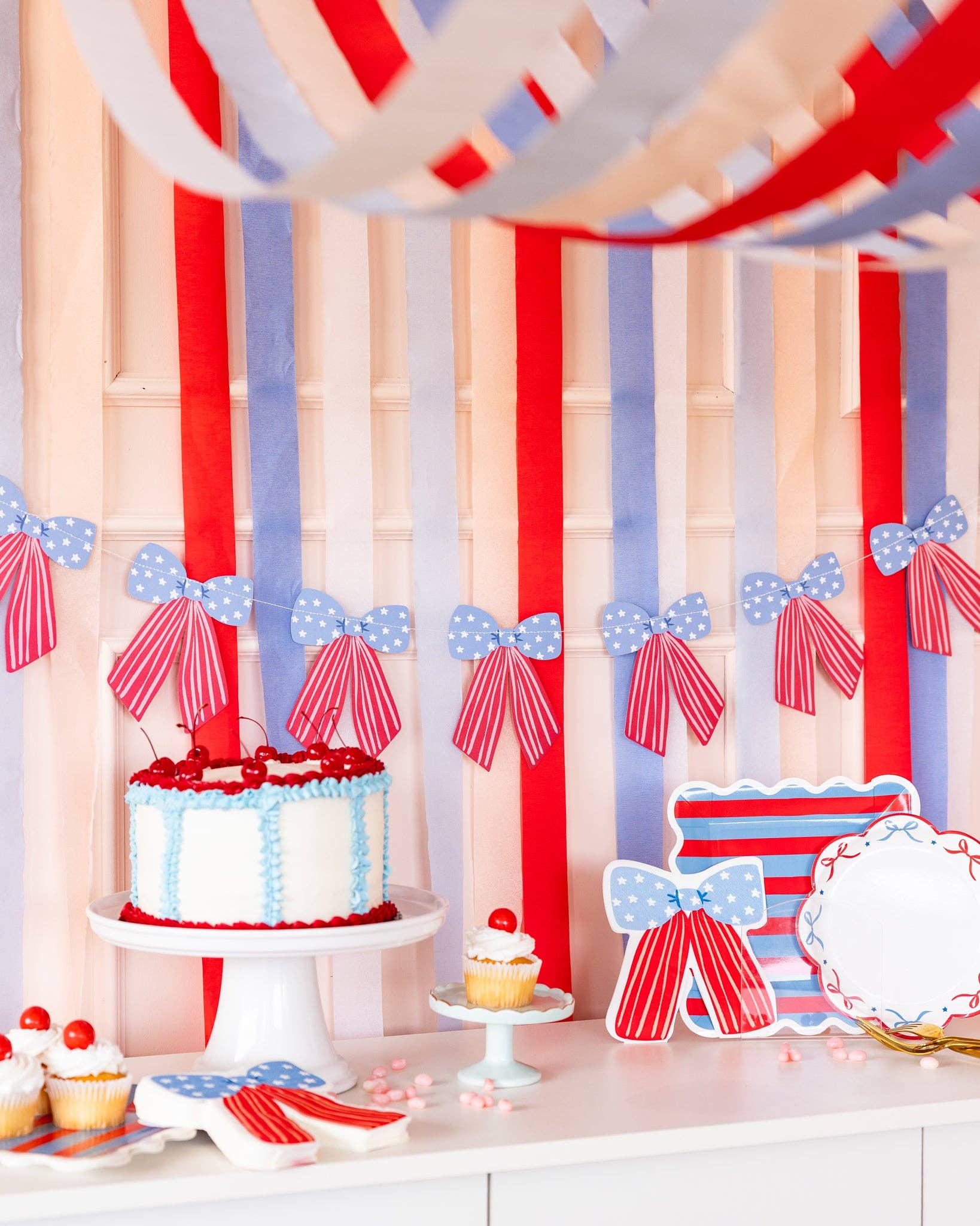 Close-up of patriotic dessert table with stars and stripes bow banner hanging over cake stand, bow-shaped plates, and striped paper plates