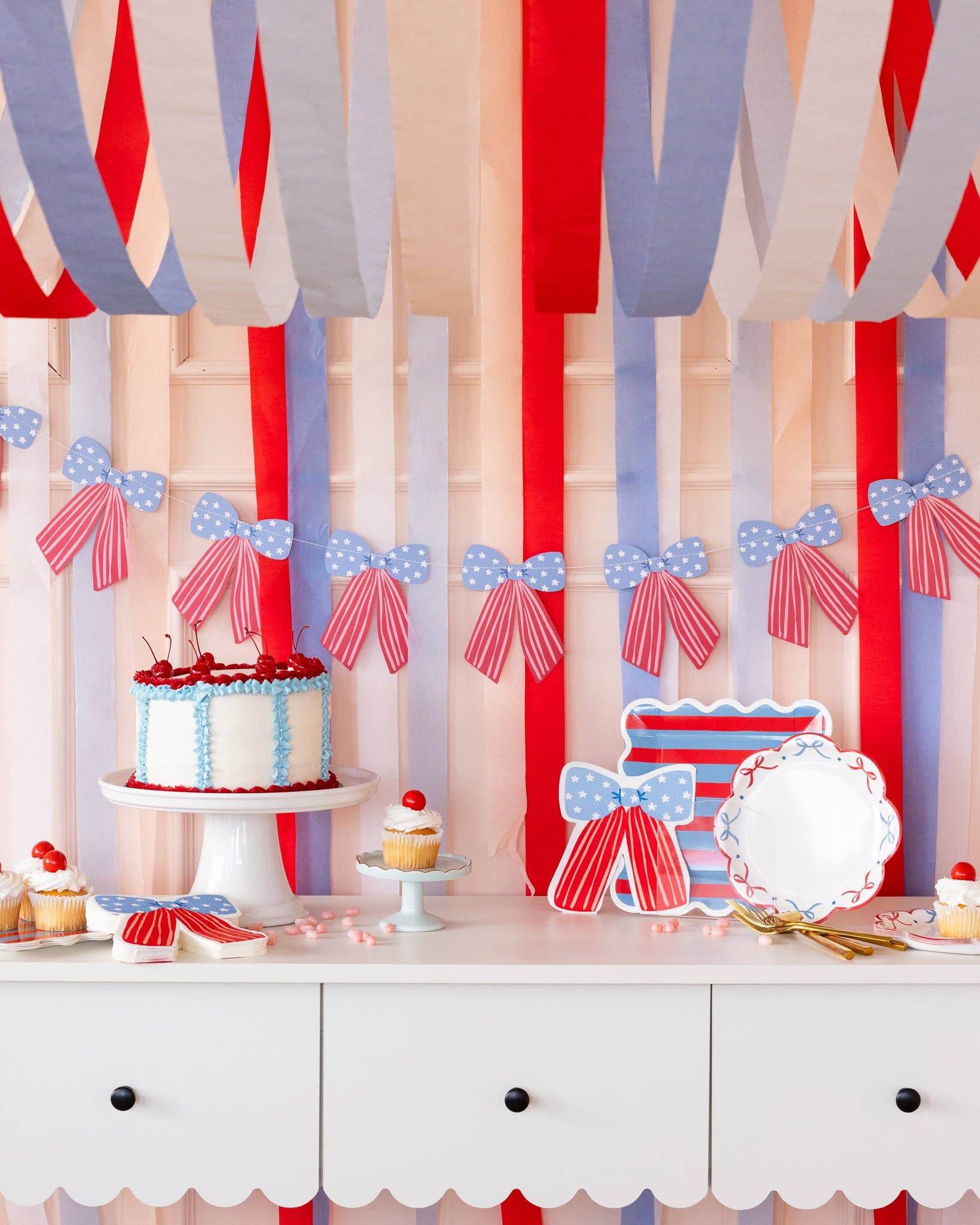 Full dessert table styled with stars and stripes bow banner, red white and blue cake, cupcakes, bow-shaped paper plates, and crepe streamers