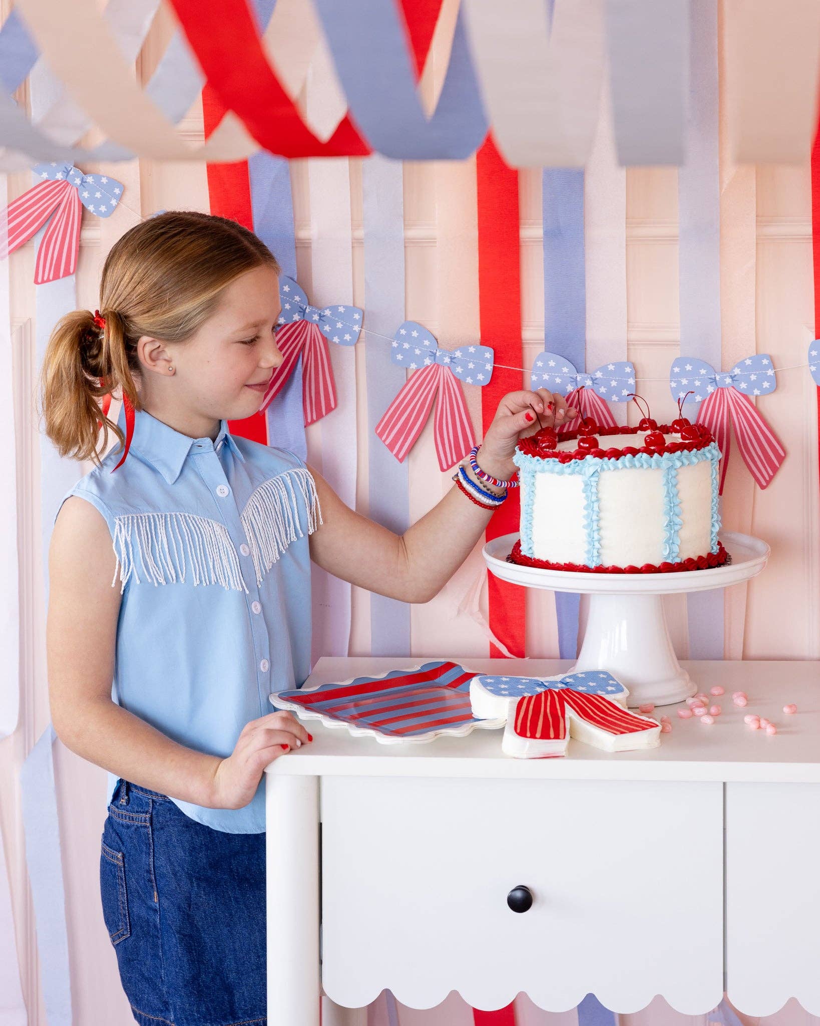 Girl decorating a cherry-topped cake at a patriotic dessert table with a stars and stripes bow banner and red white and blue streamers in the background