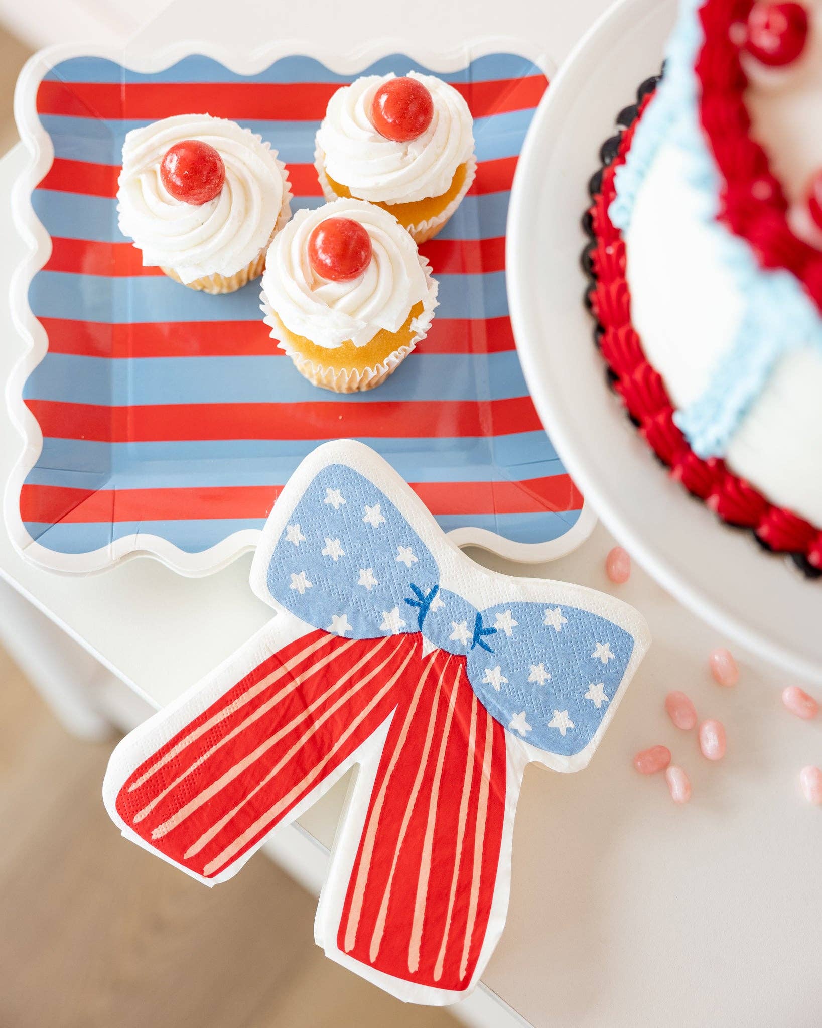 Overhead view of a stars and stripes bow shaped napkin next to cherry-topped cupcakes on a red and blue striped paper plate