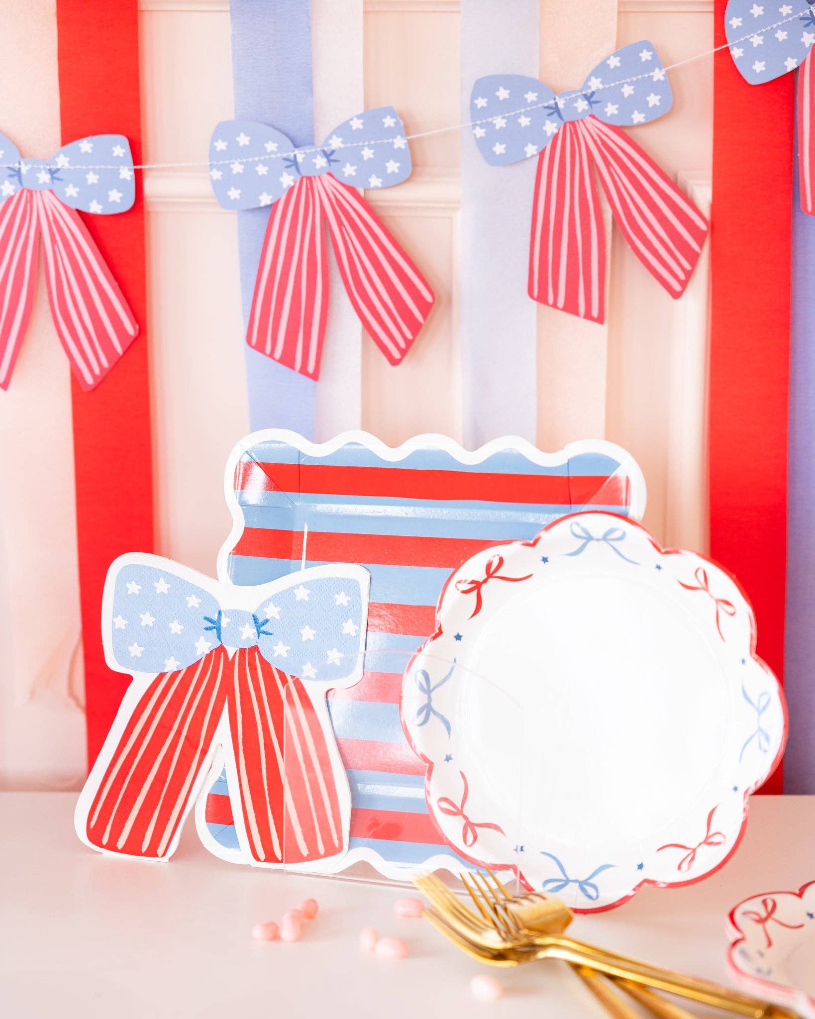 Stars and stripes bow shaped napkin displayed with matching striped paper plates and bow scalloped plates against red white and blue streamer backdrop