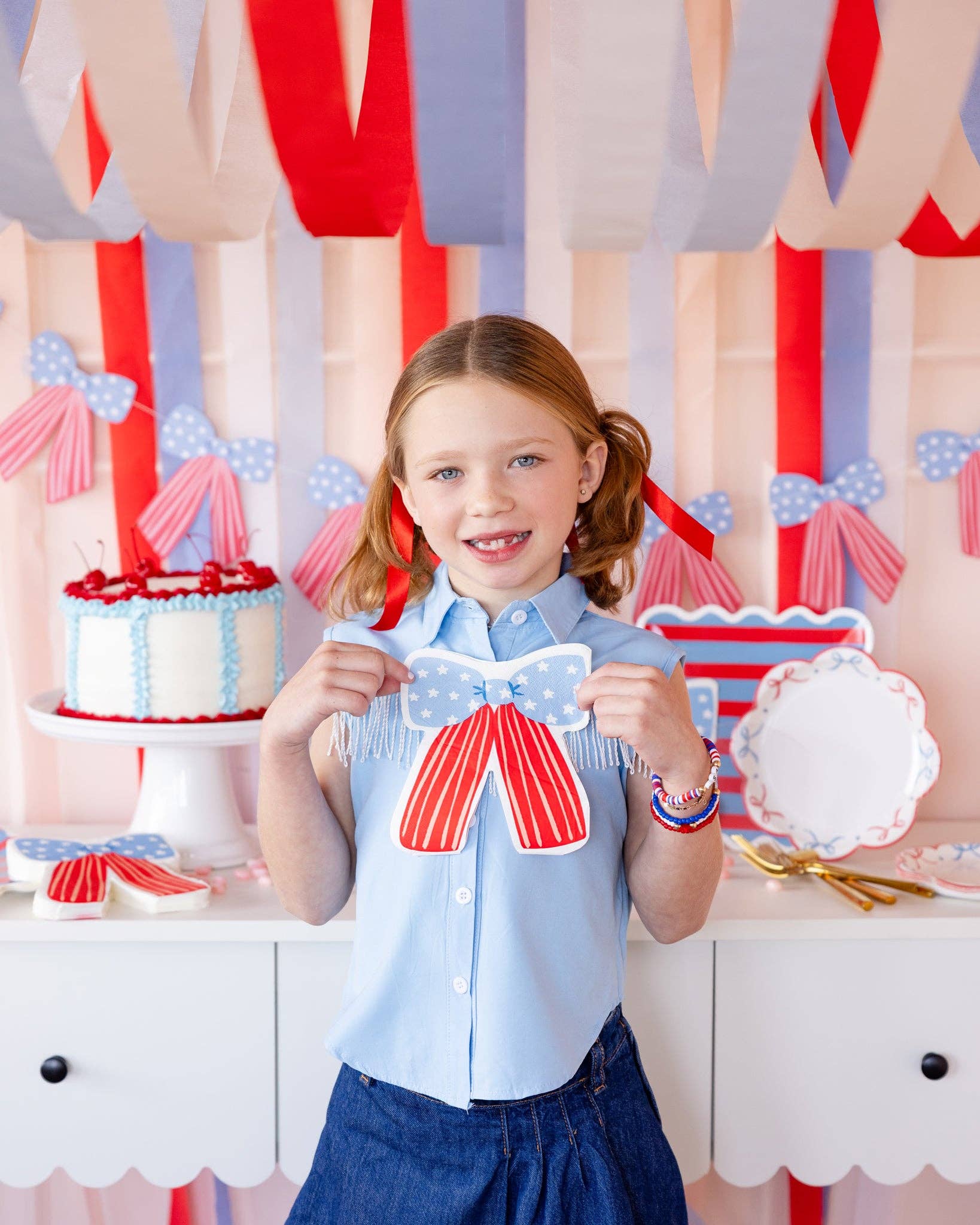 Girl holding a stars and stripes bow shaped paper napkin at a patriotic dessert table with bow banner, streamers, and red white and blue cake