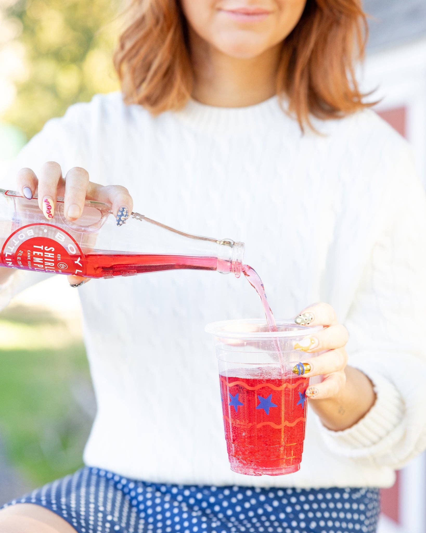 Woman pouring a red Shirley Temple into a clear plastic party cup printed with blue stars and red wavy stripes