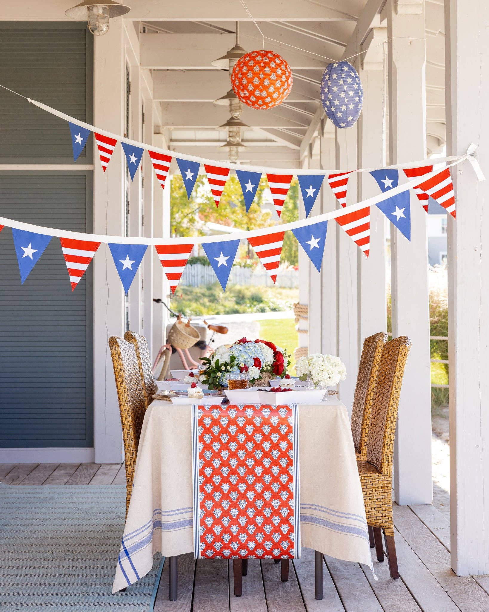 Close-up of stars and stripes pennant banner draped across porch with patriotic table setting, hydrangeas, and printed paper lanterns visible below