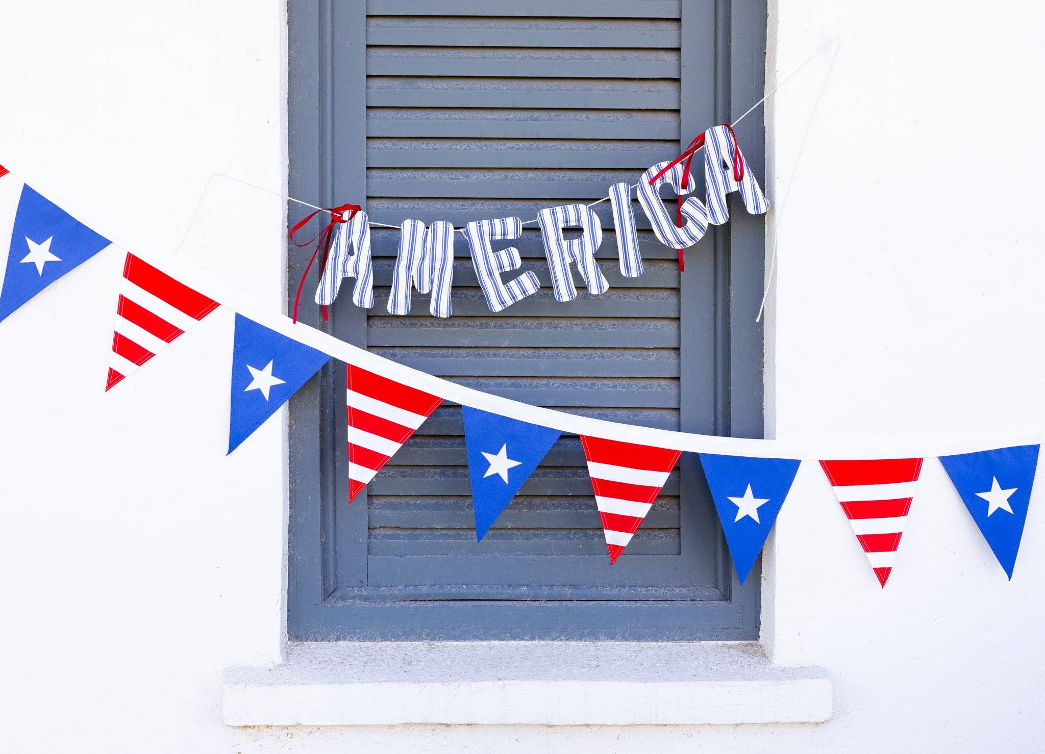 Stars and stripes pennant banner hung on exterior window shutters alongside a blue and white striped America letter banner