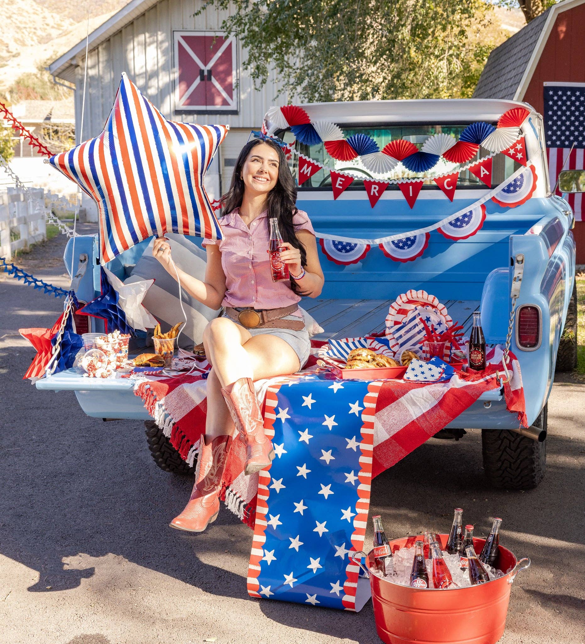 Woman sitting in a decorated blue vintage truck bed with patriotic party supplies including stars and stripes scalloped paper napkins and plates