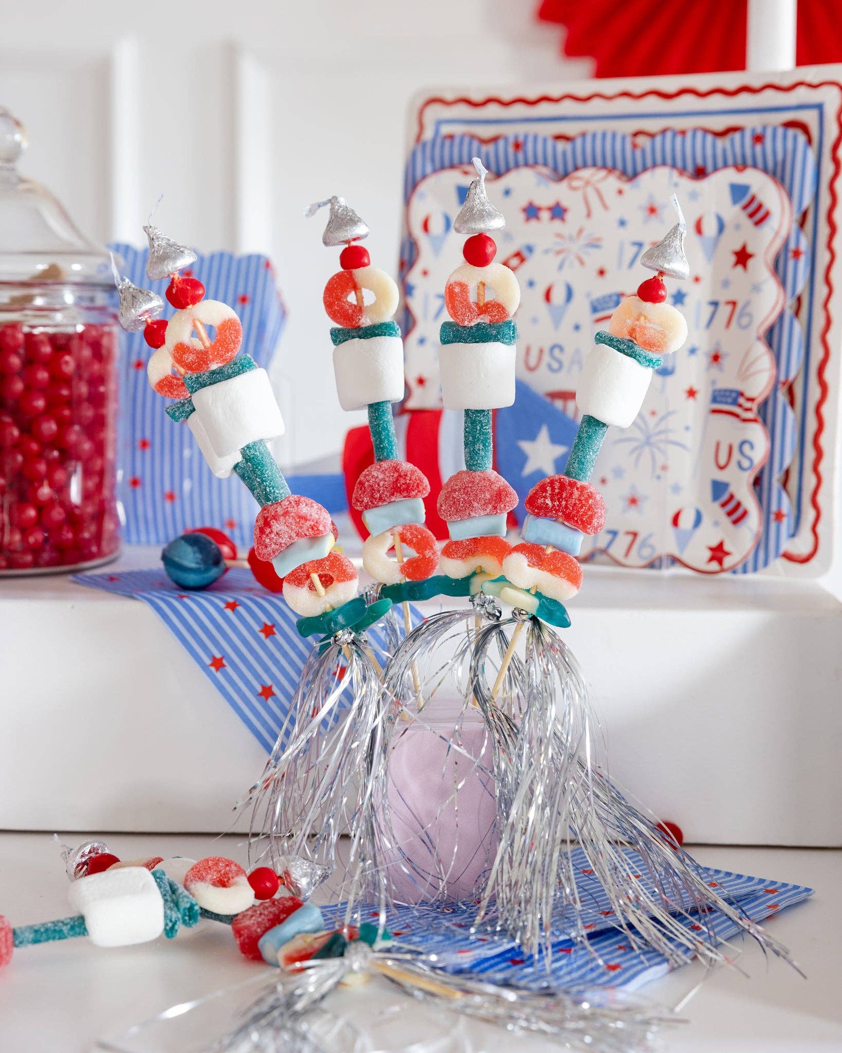 Styled Fourth of July party scene featuring stars and stripes guest napkins beneath candy kabobs with coordinating Americana plates and decor in background