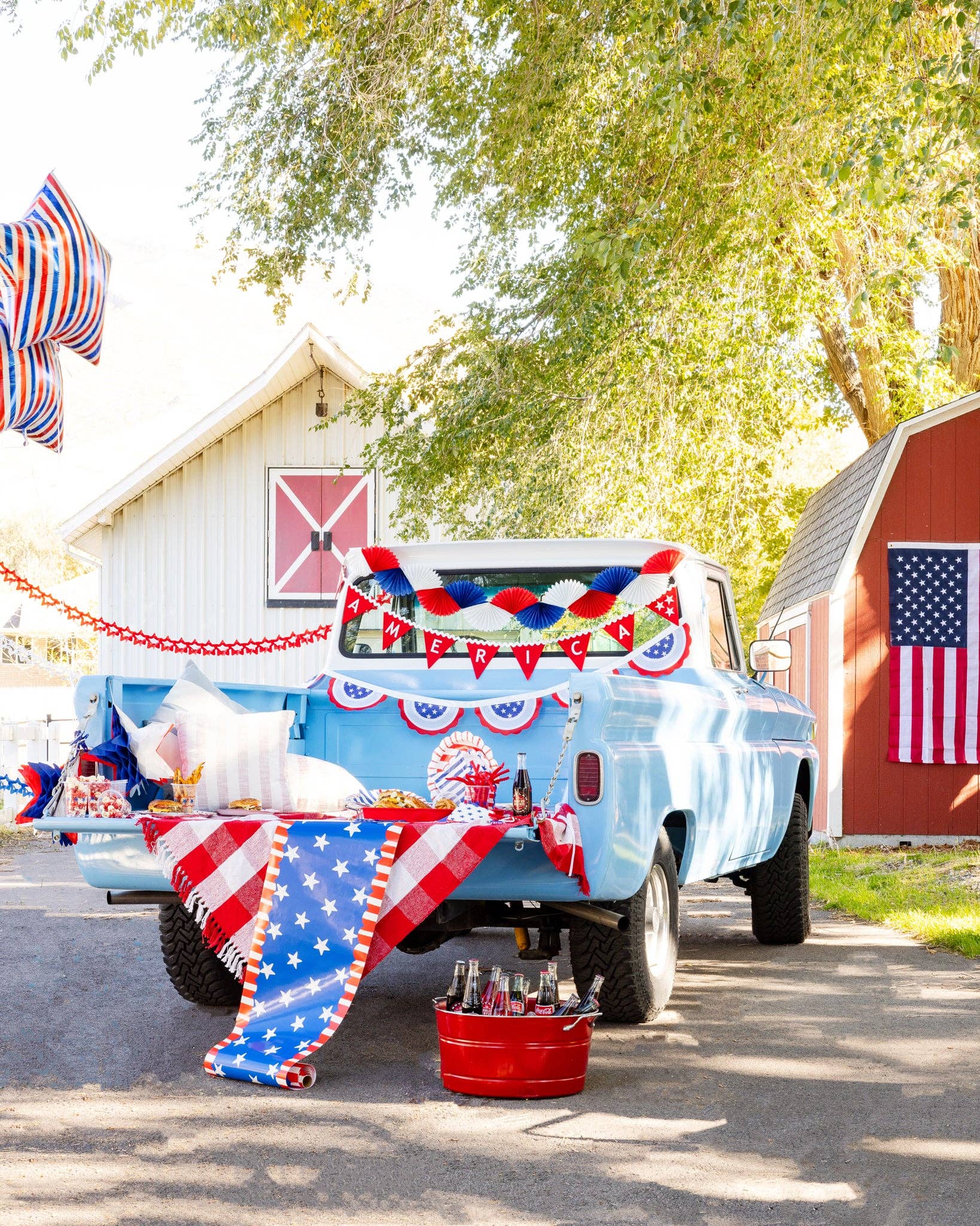 Wide shot of a patriotic truck bed party with stars and stripes paper table runner draped over the tailgate alongside coordinating plates, banners, and a red drink bucket