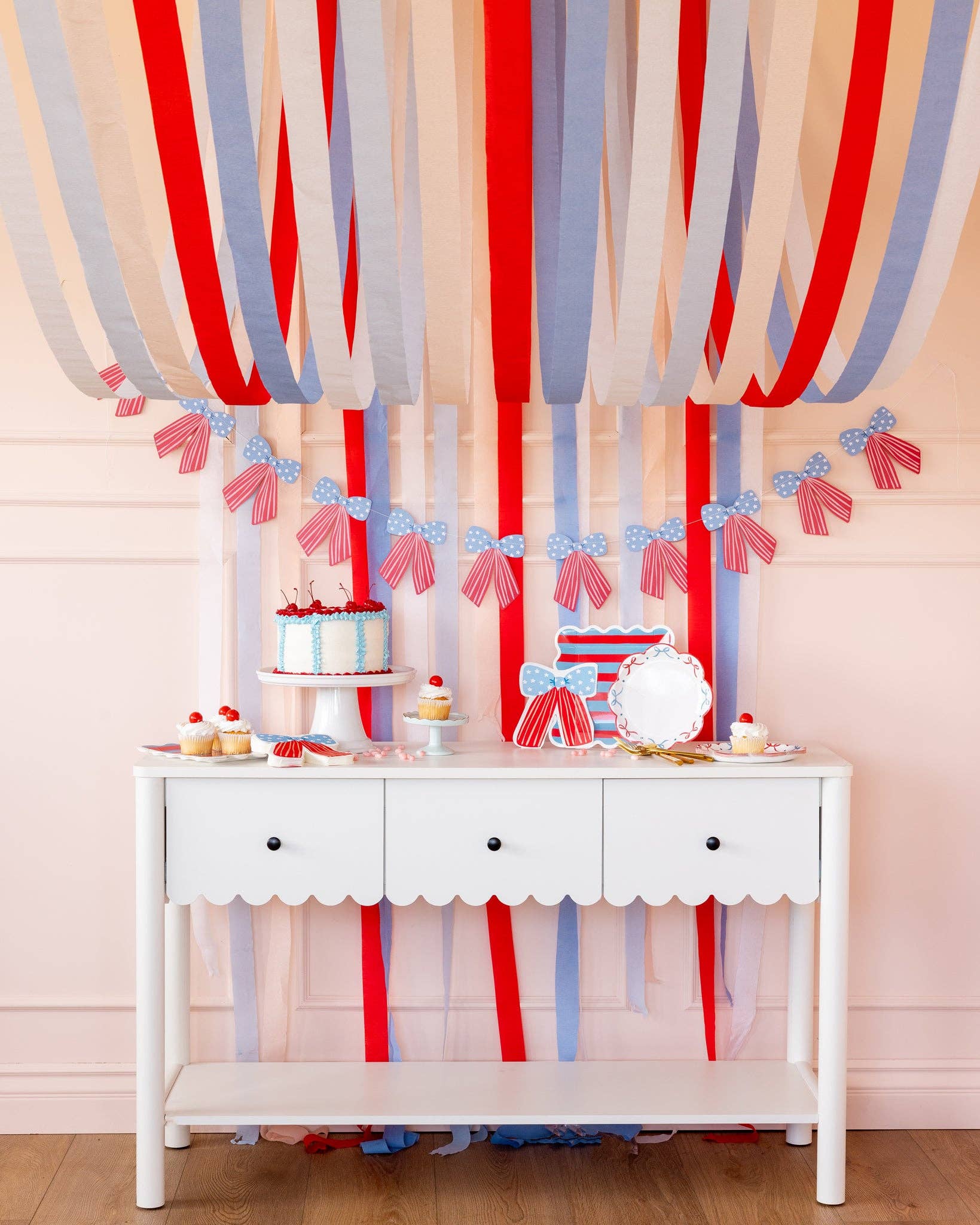 Full patriotic dessert table with red white and blue streamers, bow banner, bow shaped napkins, striped plates, and bow scalloped plates with cake and cupcakes