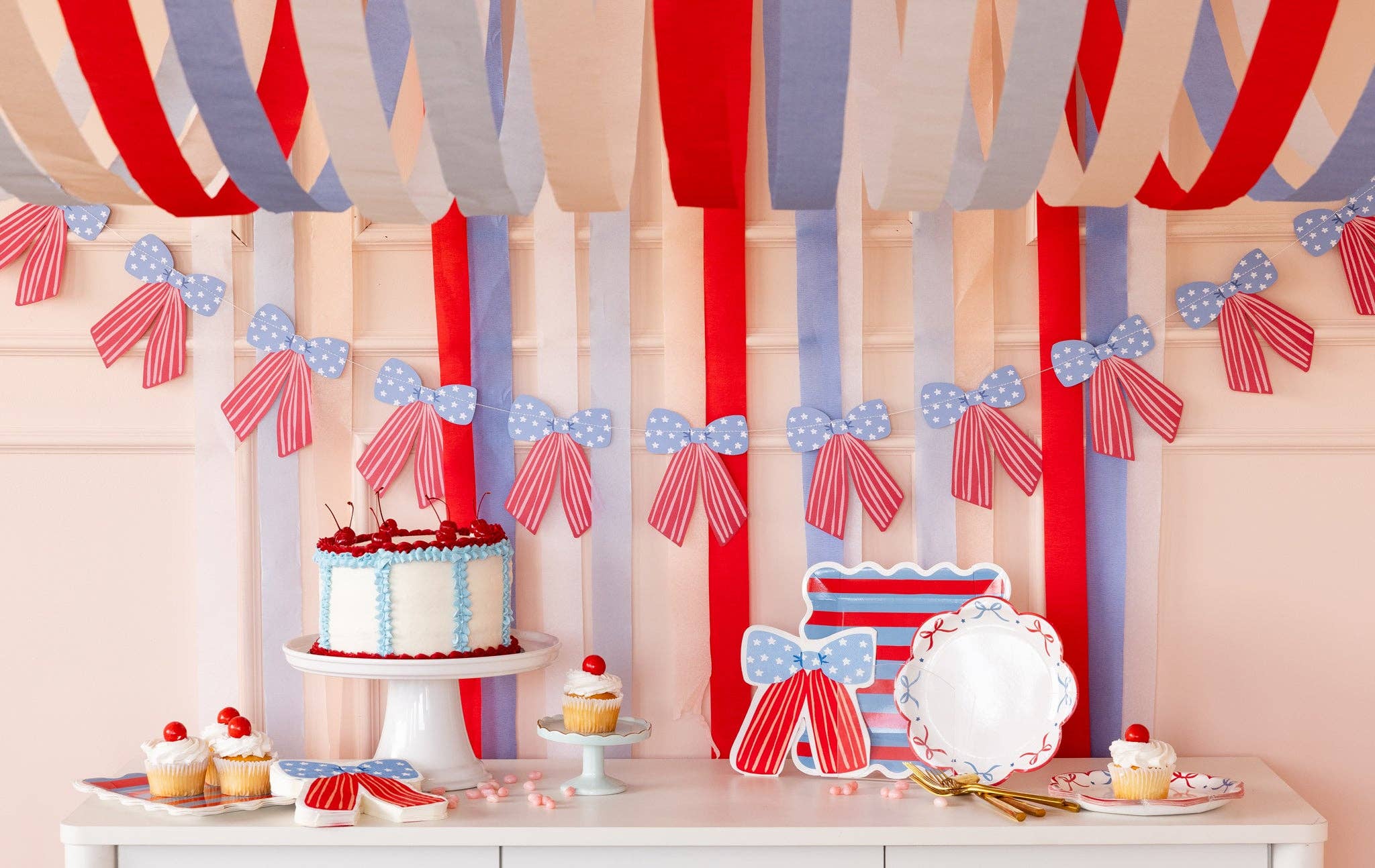 Wide view of patriotic dessert table styled with red white and blue streamers, bow banner, bow shaped napkins, striped plates, bow plates, cake, and cupcakes