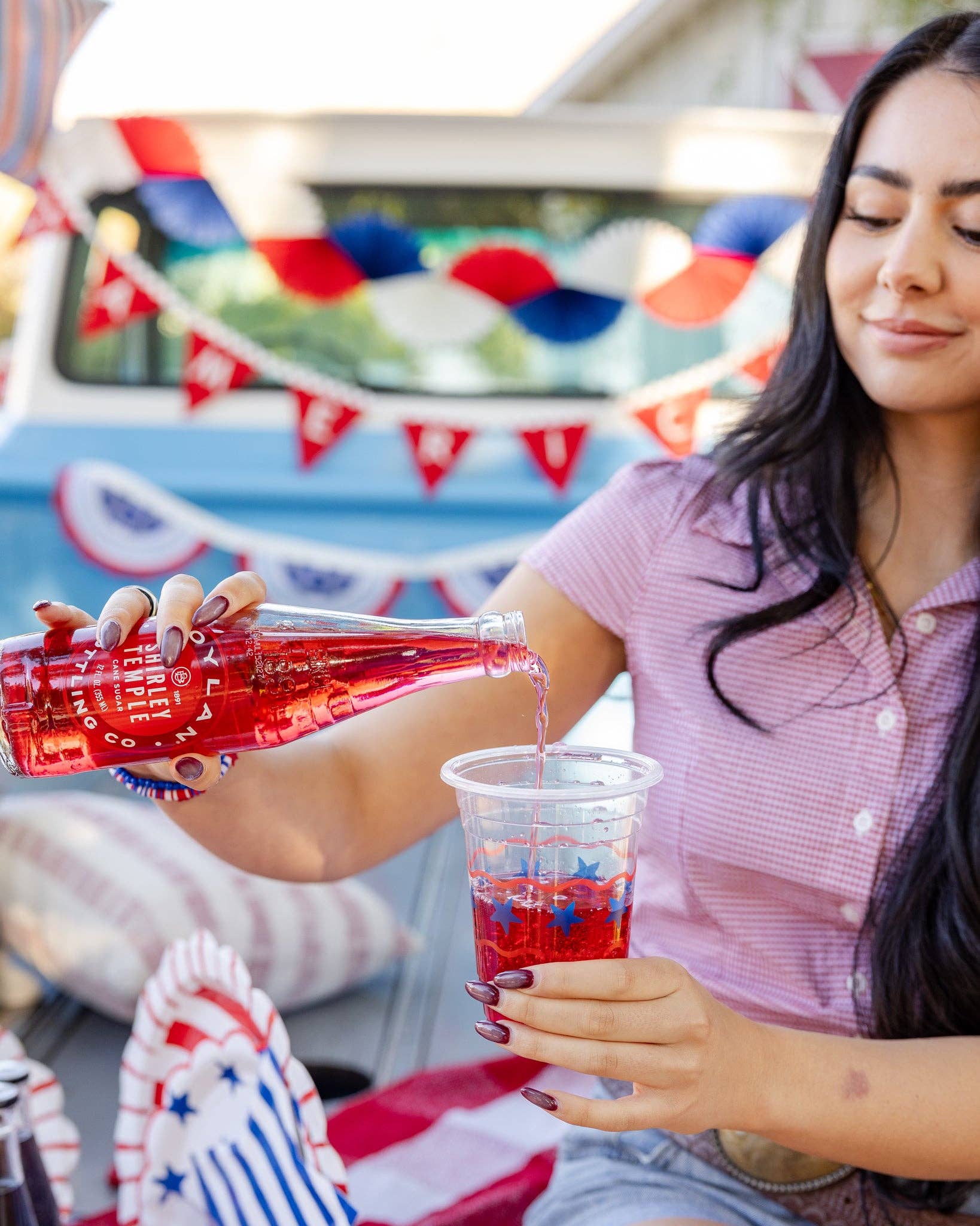 Woman pouring a red drink into a stars and stripes clear plastic party cup with patriotic decorations in the background