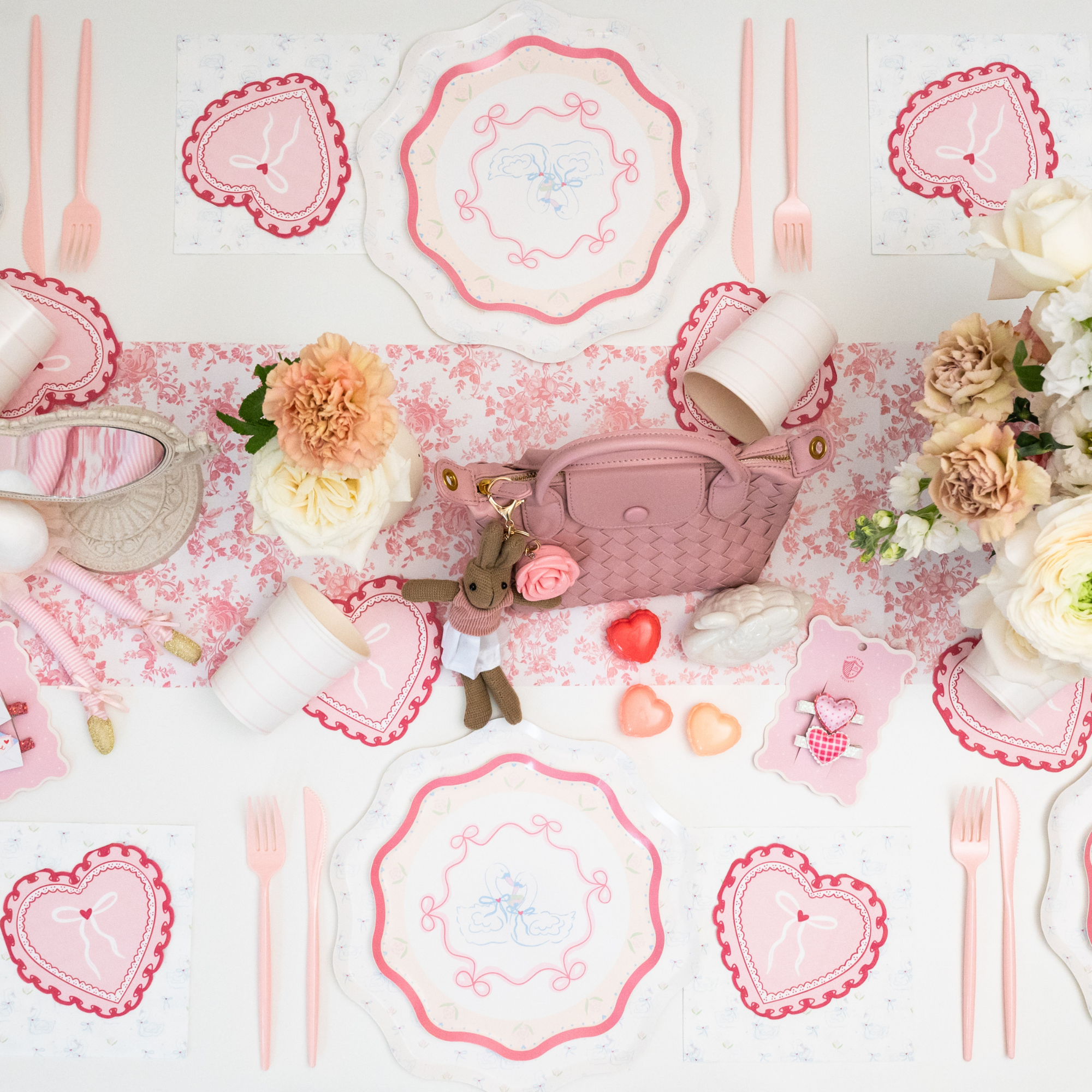 Overhead view of coquette Valentine's Day party table with swan love birds scalloped plates, heart shaped napkins, pink cutlery and floral table runner with roses and carnations