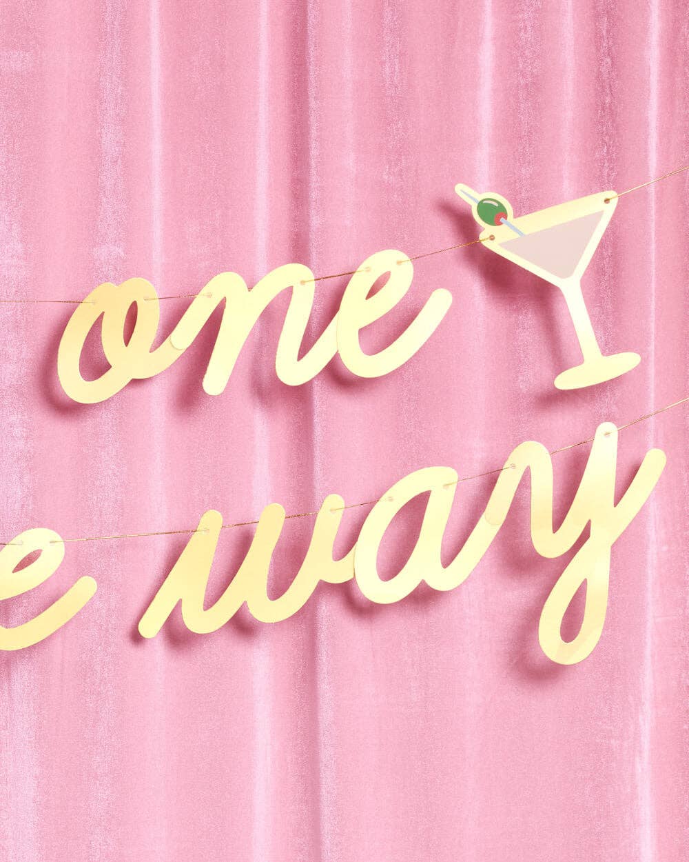 Close-up of gold foil script and martini glass icon on Tini One on the Way banner against pink velvet
