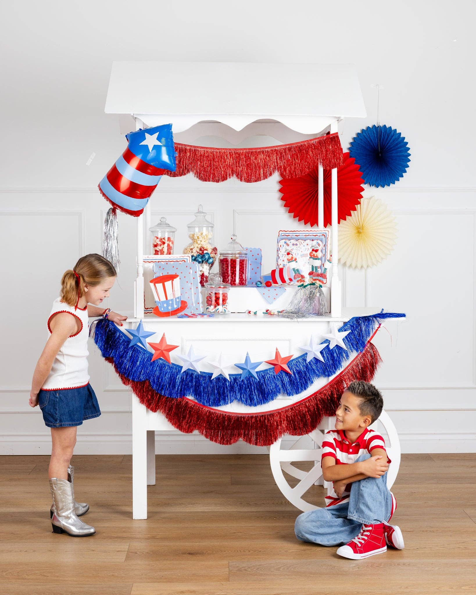 Patriotic party cart decorated with red, white, and blue supplies featuring Uncle Sam hat shaped paper plates, star garlands, tinsel fringe, and candy displays with two children