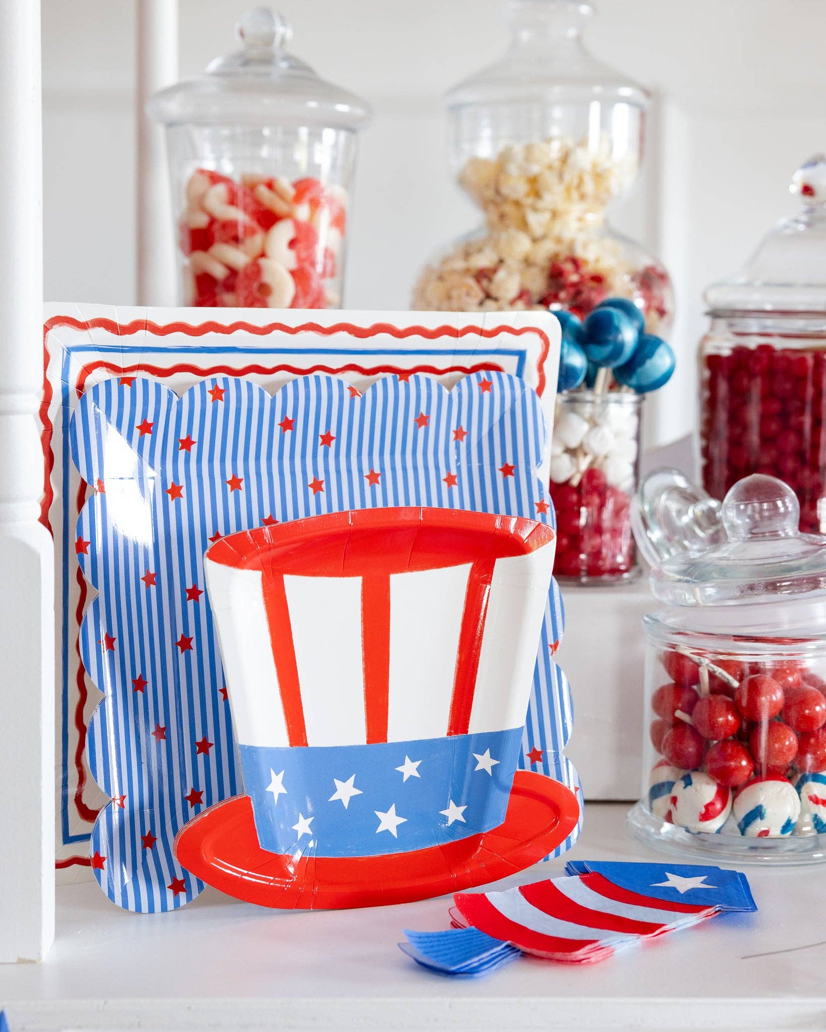 Uncle Sam hat shaped paper plate displayed in front of stars and stripes scalloped plates on a patriotic dessert table with glass candy jar