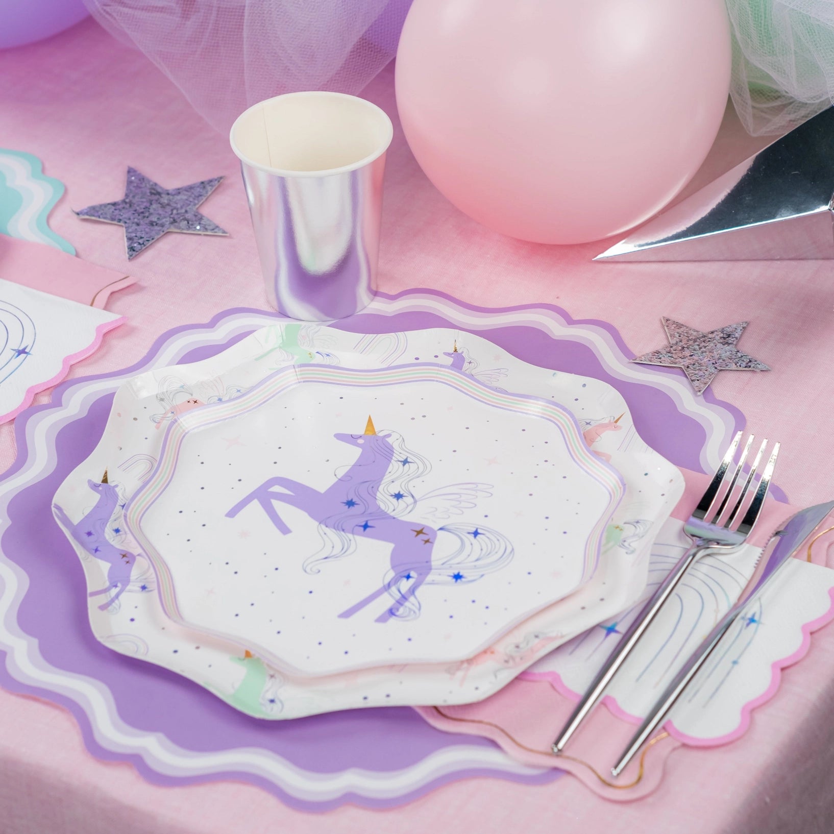 Unicorn-themed paper plates with cutlery on a pink surface with balloons and stars.