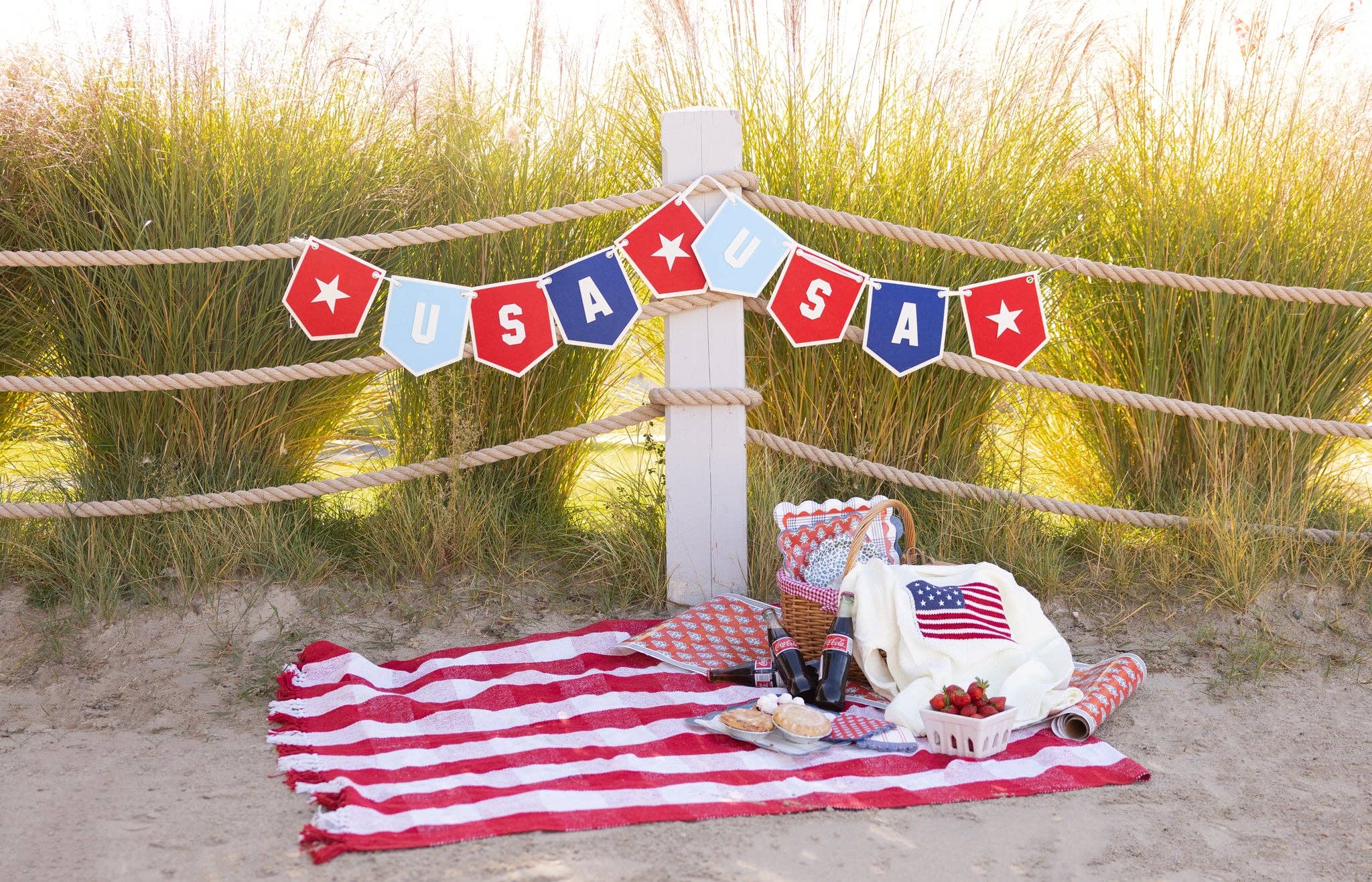 Wide shot of USA felt pennant banner displayed on beach rope fence at golden hour with American flag picnic blanket and basket below