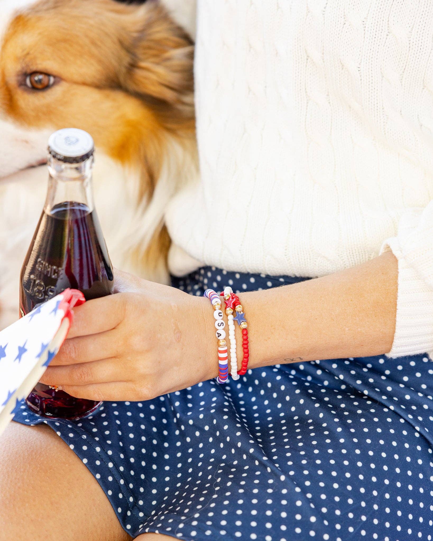 Close-up of a woman's wrist wearing three patriotic beaded bracelets with USA letter beads, star charms, and red white and blue seed beads while holding a soda bottle