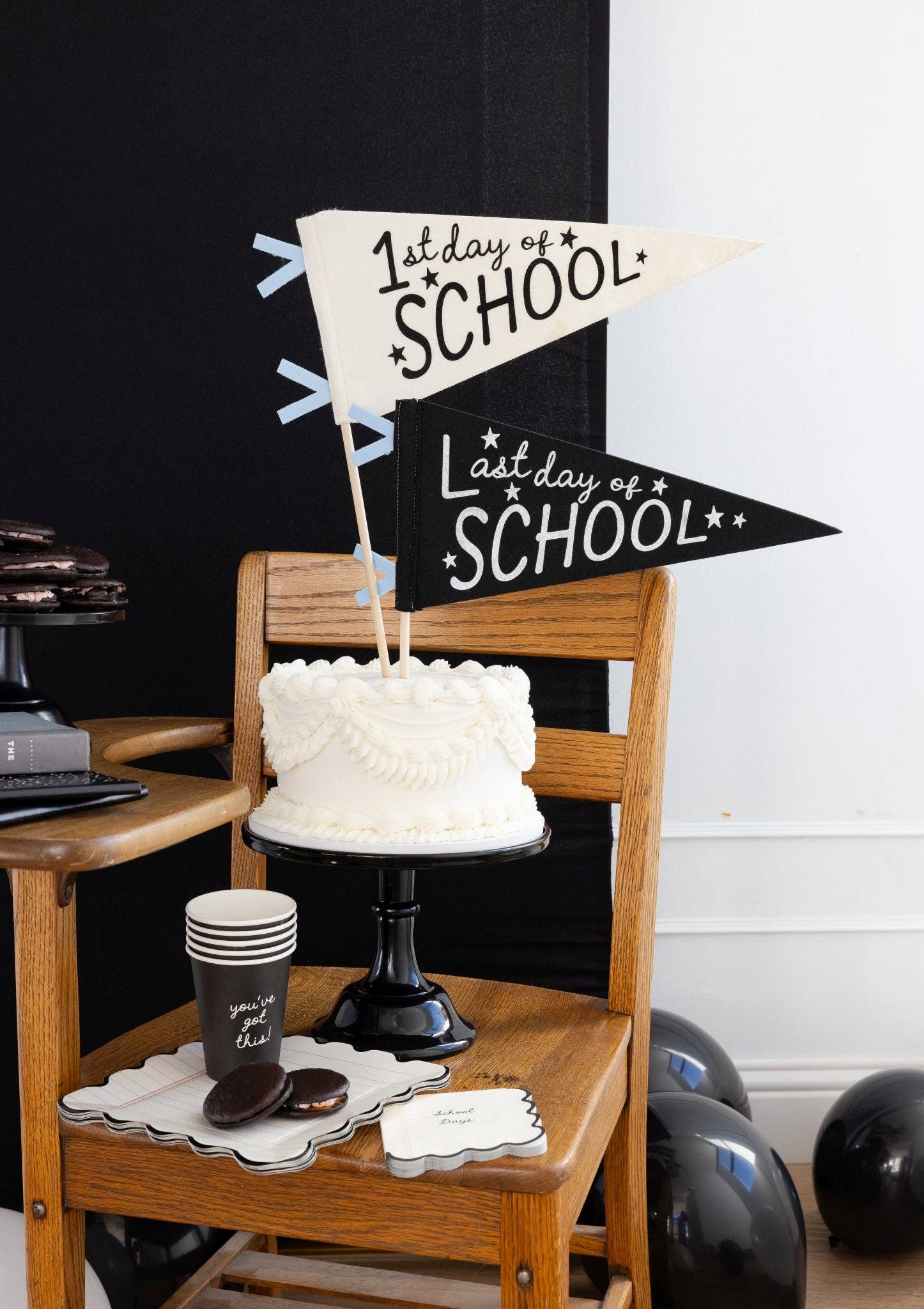 Styled back to school dessert scene with black "you've got this" paper cup next to chocolate whoopie pies on a notebook paper scalloped plate, with white cake on a black cake stand and 1st Day and Last Day of School pennants